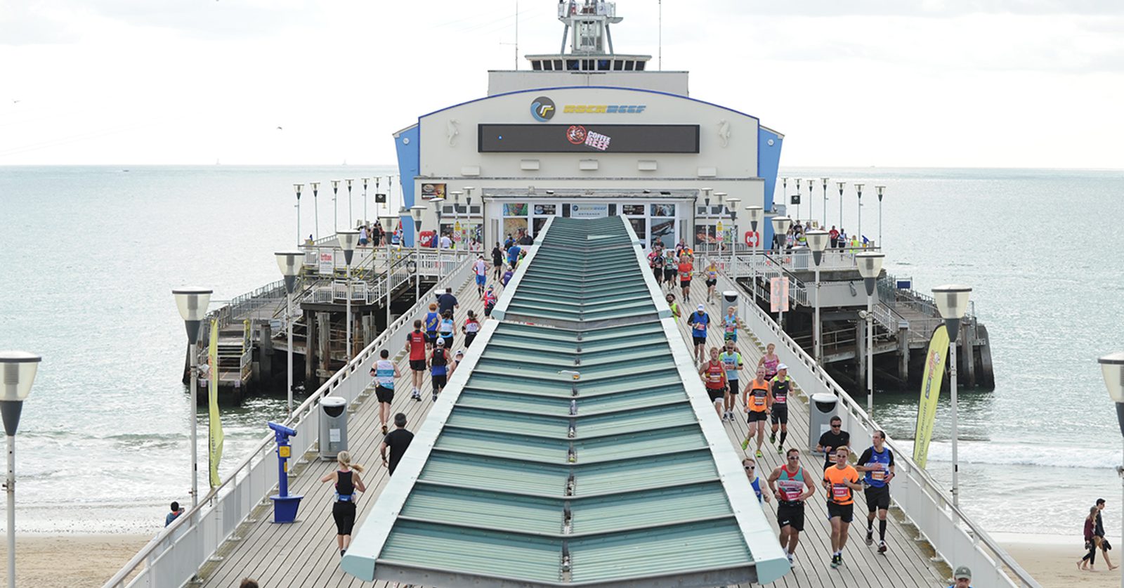 Participants run along a pier towards the ocean, taking part in a race event. The pier stretches out into calm water, and a large building is visible at the end. The sky is overcast, and some spectators are standing along the sides, watching the runners.