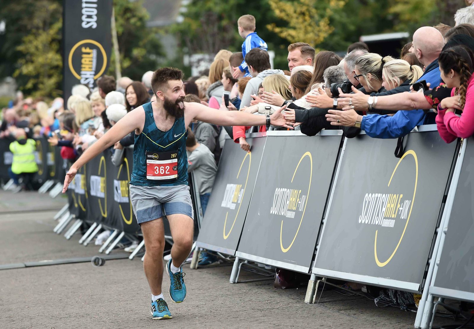 A male runner with a race bib numbered 362 high-fives spectators lined along the race route, who are cheering and reaching out. The crowd is gathered behind barriers with banners. The runner is wearing a teal tank top, grey shorts, and blue running shoes.