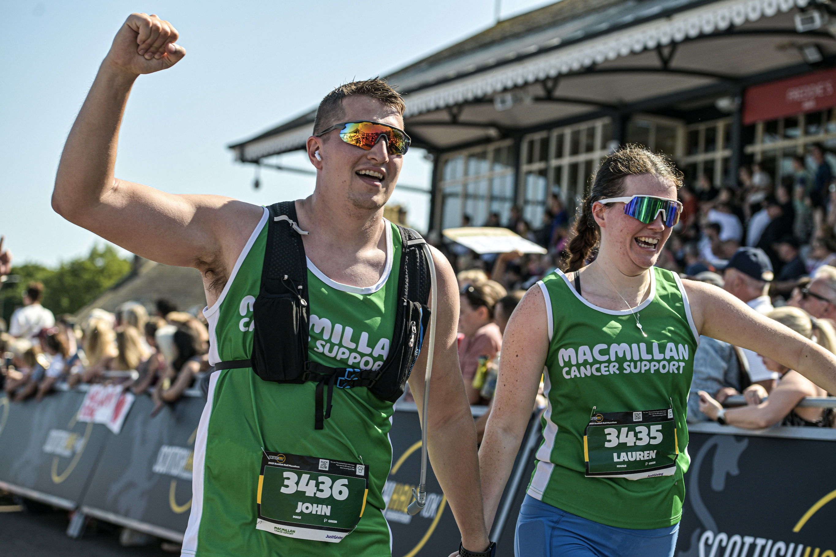 Two marathon runners wearing green Macmillan Cancer Support vests celebrate at the finish line. Both are smiling, with one raising a fist in triumph. A cheering crowd and building are visible in the background.