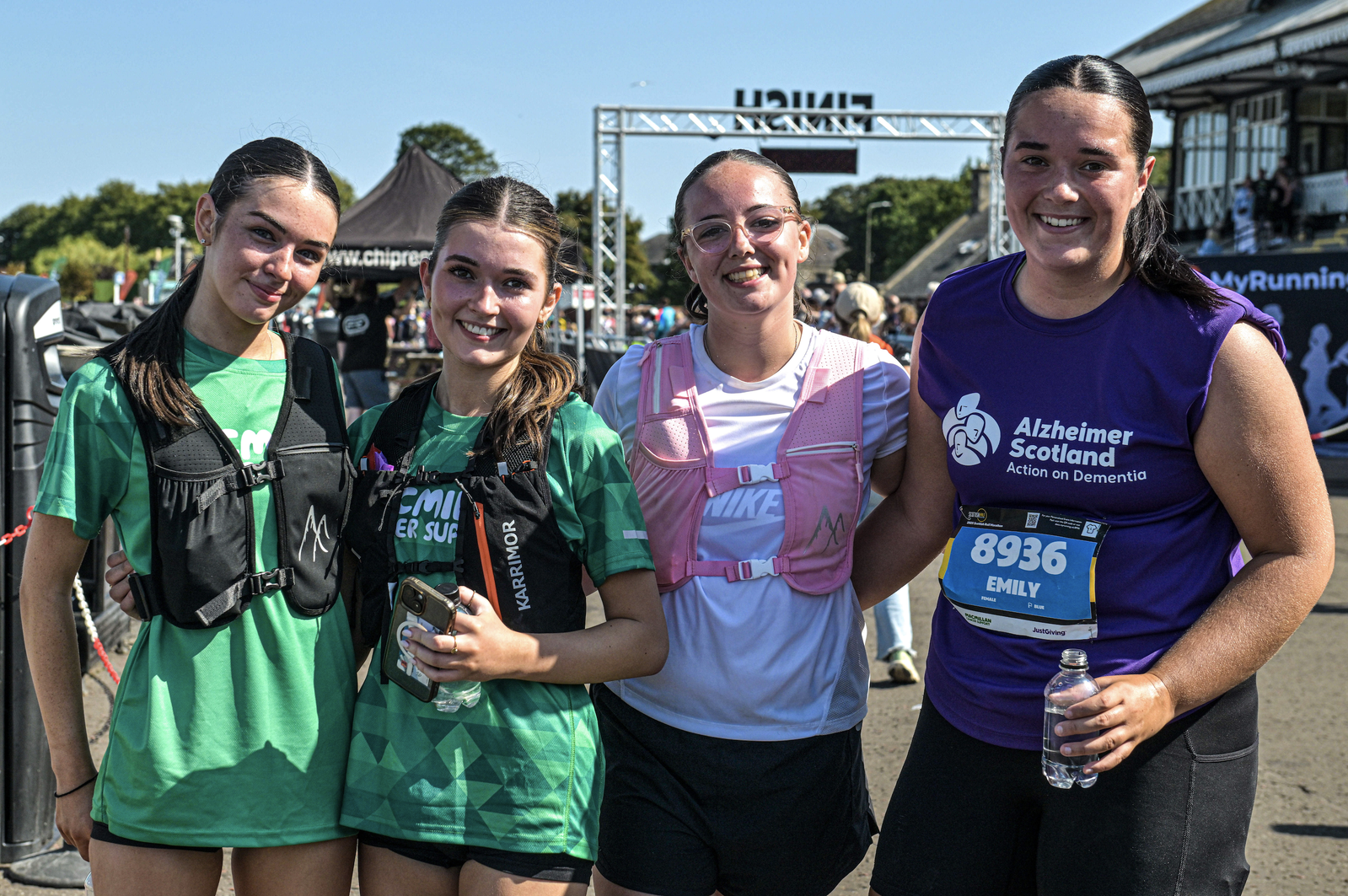 Four young women wearing athletic gear smile and pose together after a race. One wears a purple Alzheimer Scotland shirt and a race bib marked "Emily 8936." Sunny weather and event signage are visible in the background.