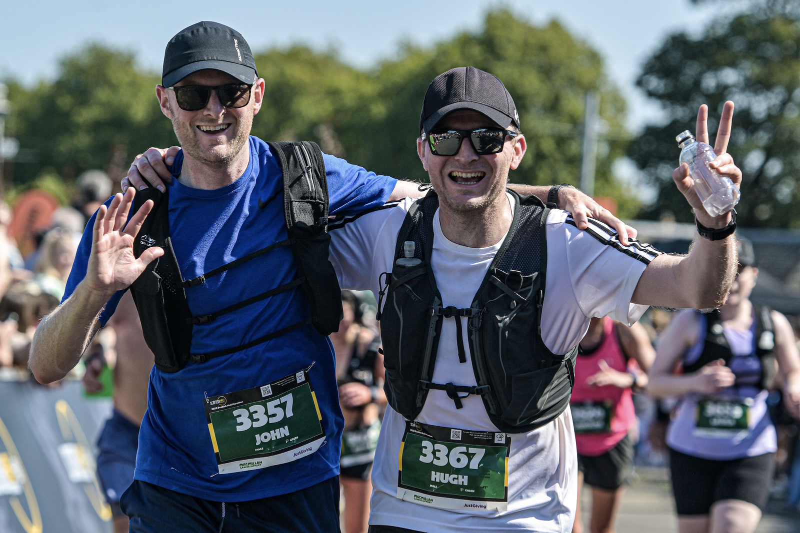 Two male marathon runners smiling and waving at the camera, wearing hats, sunglasses, and race bibs with the names John and Hugh, surrounded by other runners on a sunny day.
