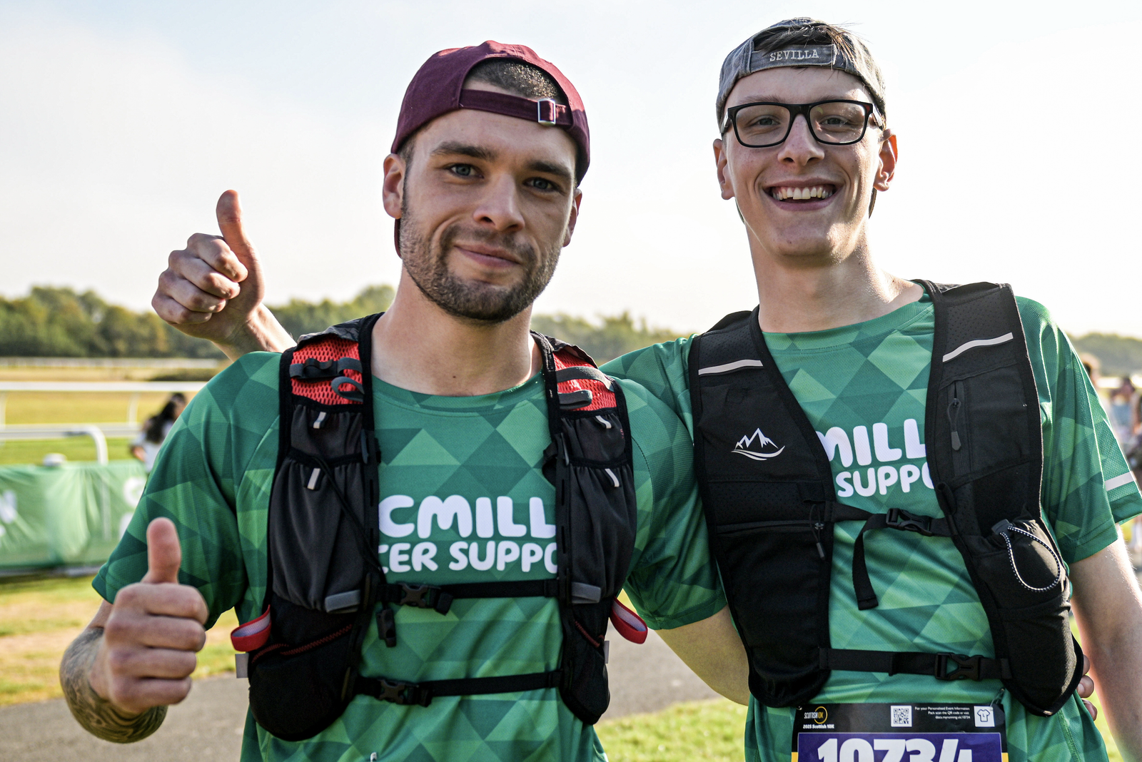Two smiling runners in green "Macmillan Cancer Support" shirts stand side by side, each giving a thumbs up. They wear hydration packs and race bibs, posing outdoors on a sunny day.