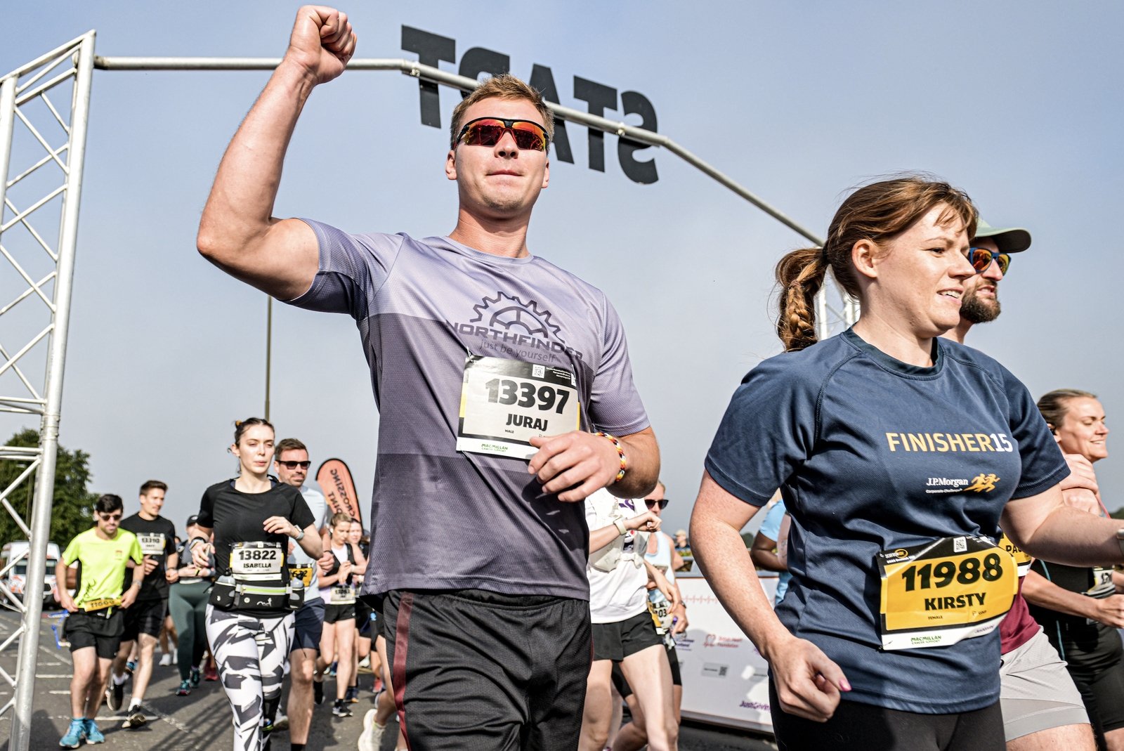 Runners at a race start line, including a man raising his fist and wearing a numbered bib, and a woman in a “Finishers” shirt, with others running behind them on a sunny day.