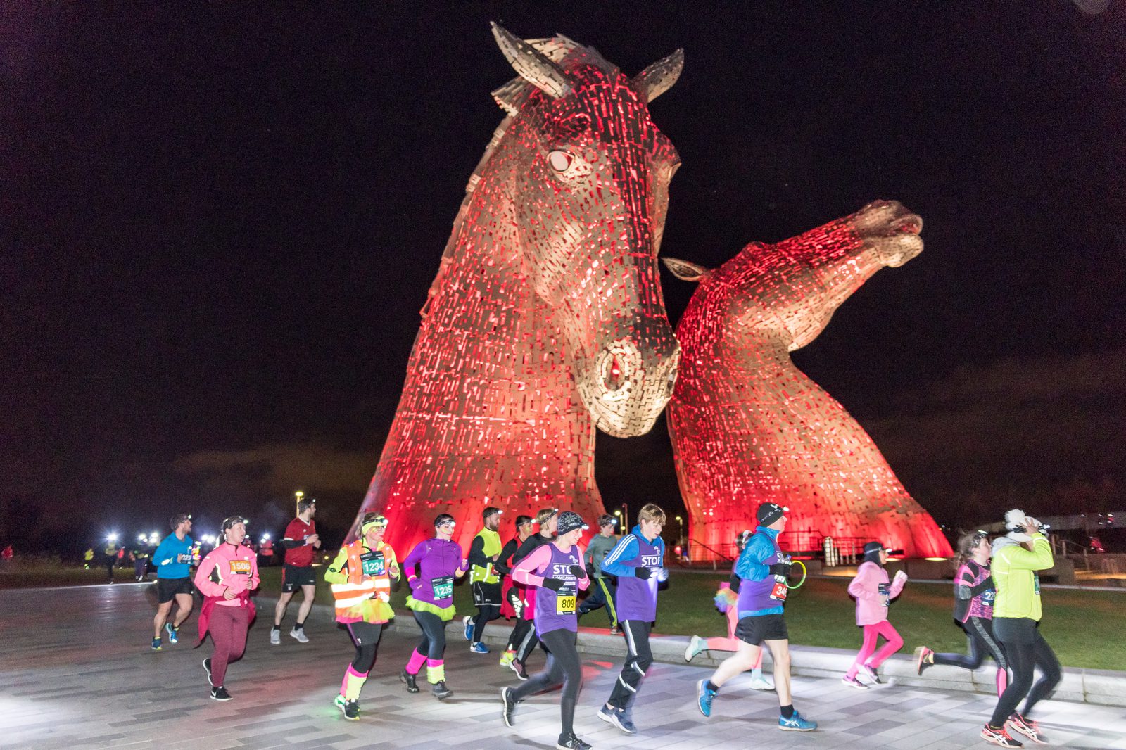 Runners participate in a night-time event in front of the Kelpies sculptures, which are large horse head statues illuminated with red lights, set against a dark sky. The participants wear colorful athletic gear.