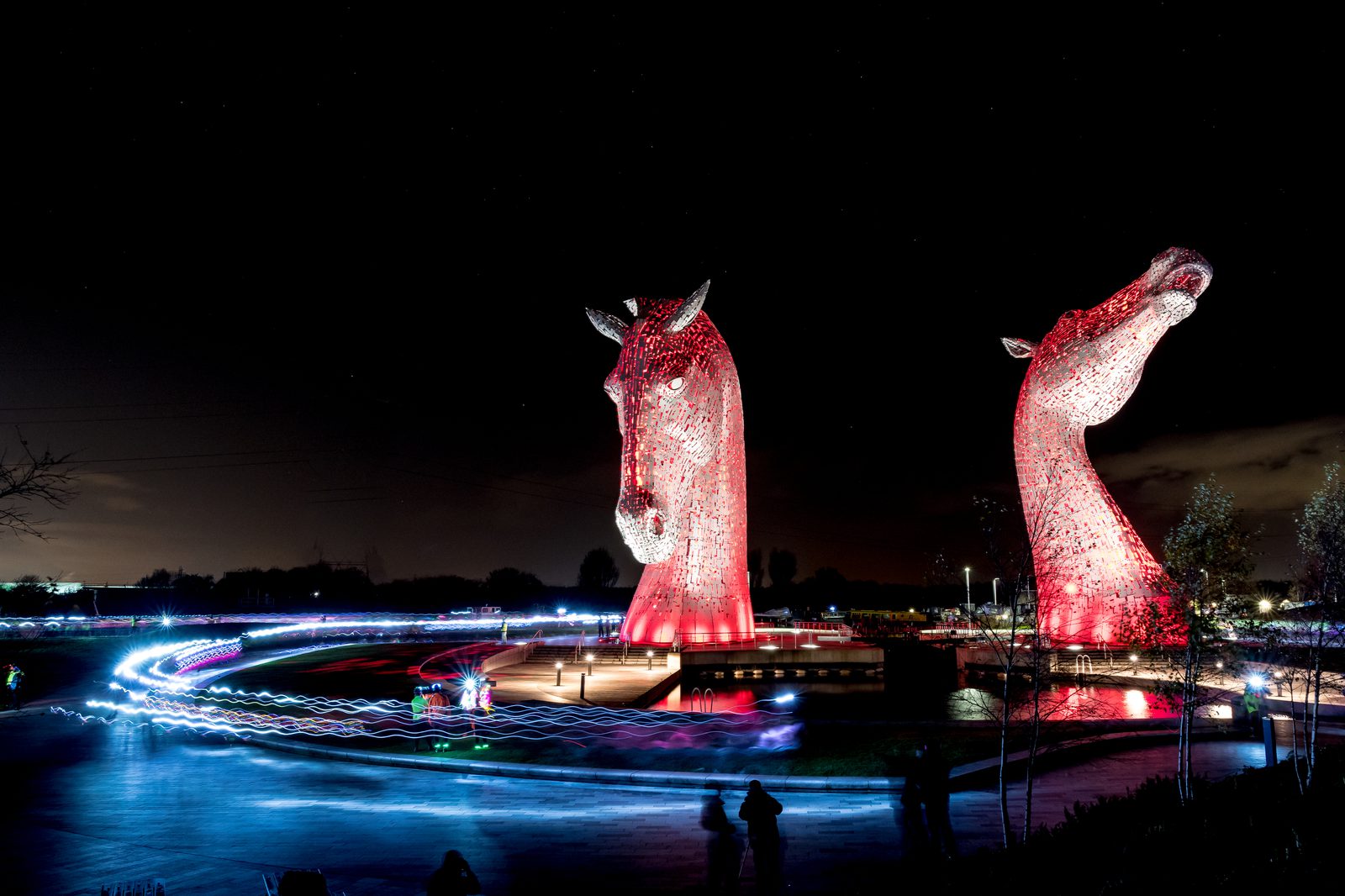 A nighttime view of The Kelpies, two large horse head sculptures illuminated in red. Light trails from passing vehicles or light sources can be seen around the sculptures, creating a dynamic effect. The sculptures are set in a park with a dark sky in the background.