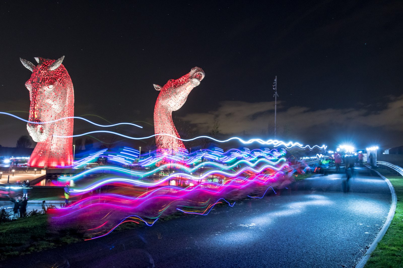 A nighttime scene featuring two large, illuminated horse head sculptures against a dark sky. The foreground is streaked with multi-colored light trails, creating a dynamic effect. The pathway is lined with onlookers observing the light display.