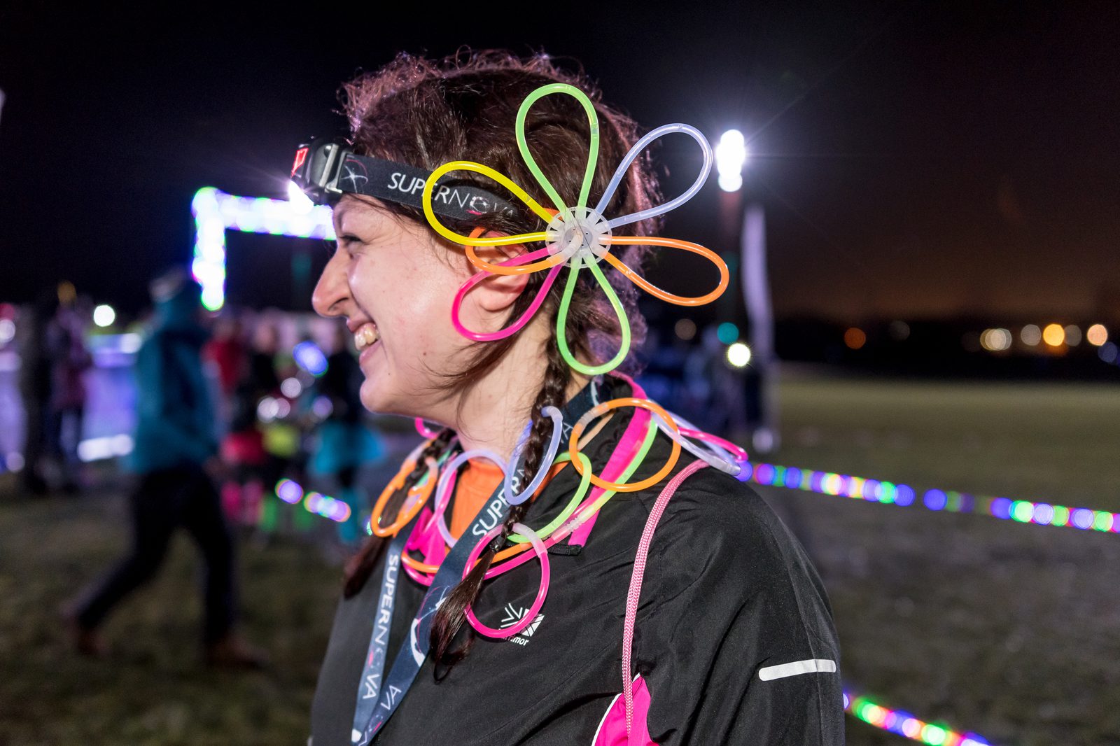 A person is smiling at night during a festive event. They wear a headband with a light on it and neon glow stick decorations shaped like flowers on their head and around their neck. People and colorful lights can be seen in the background.