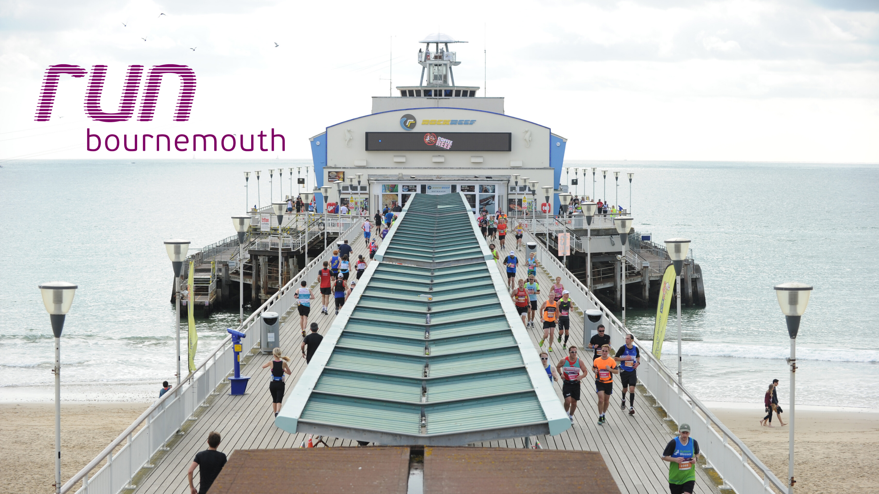Runners participate in a race on Bournemouth Pier in Bournemouth, UK. The sky is cloudy, and the ocean is calm. The event banner reads "Run Bournemouth." The pier is bustling with runners heading towards the finish line.