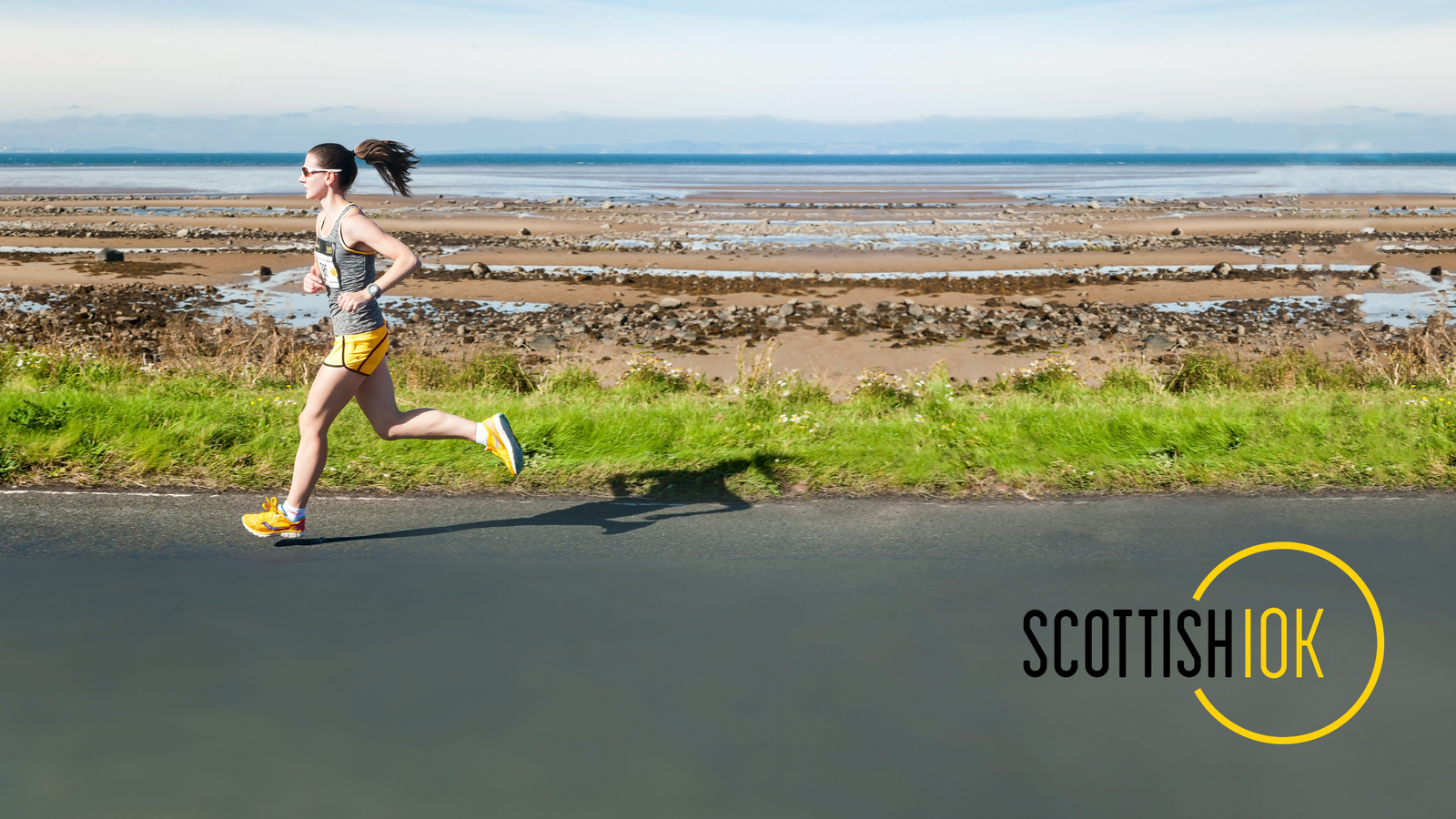 A person jogs along a paved road next to a grassy area, with a rocky shoreline and the sea in the background. The sky is clear and sunny. The text "Scottish OK" is displayed in the bottom right corner, with "OK" enclosed in a yellow circle.