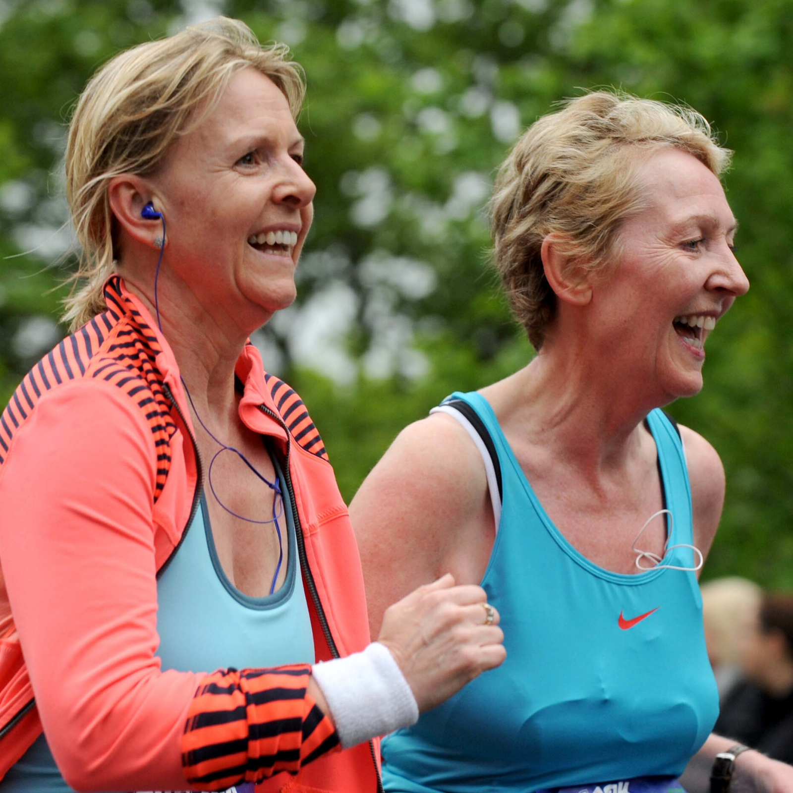 Two women in athletic wear are running outdoors, smiling joyfully. Both have earphones in. The text "WOMEN'S 10K" and a star symbol are visible on the left side of the image. The background is blurred but appears to be a green, leafy park with other runners.
