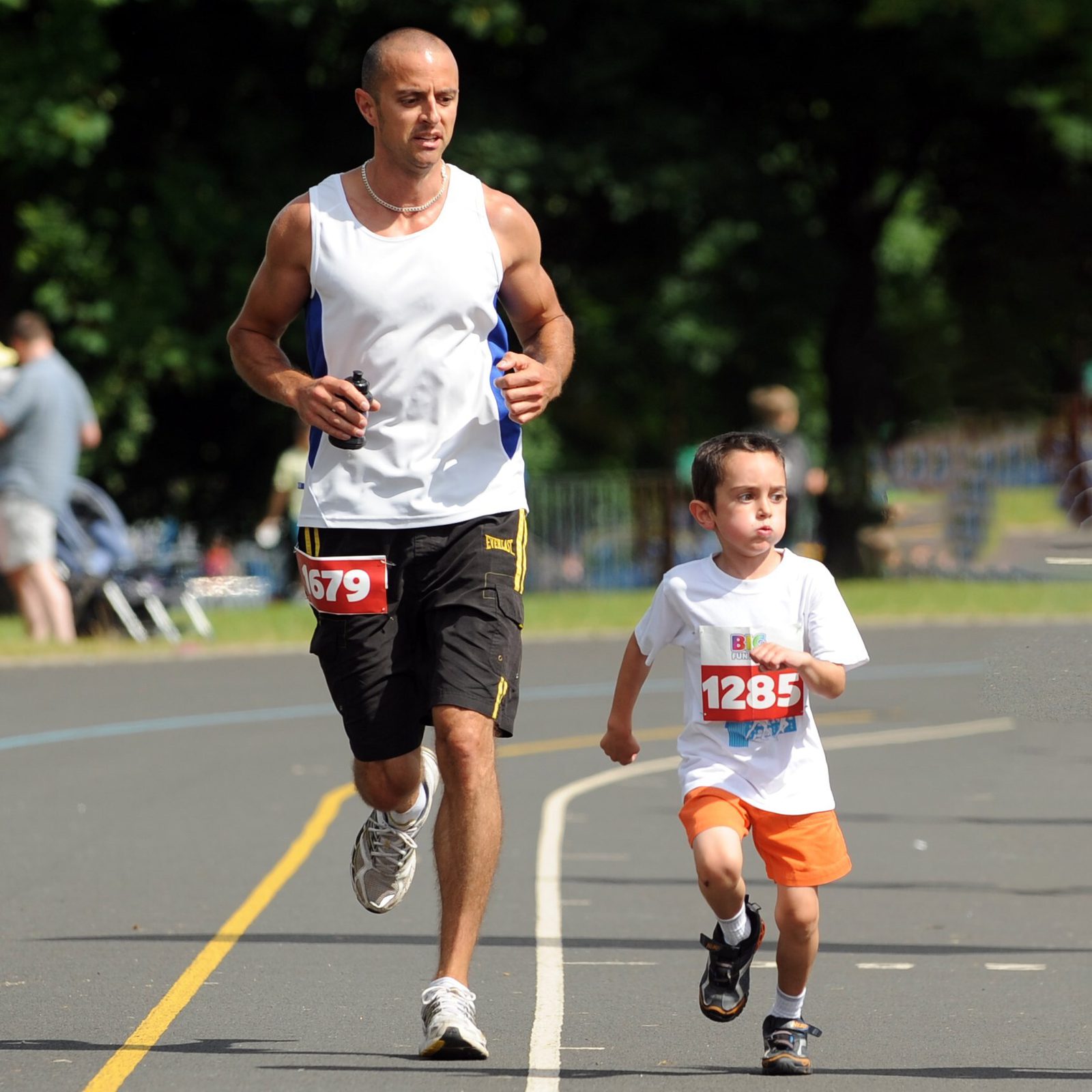 An adult and a child are running side by side on a road, both wearing race bibs. The adult is in a white tank top and black shorts, holding a water bottle, while the child is in an orange shorts and a white t-shirt. Trees and spectators are visible in the background.