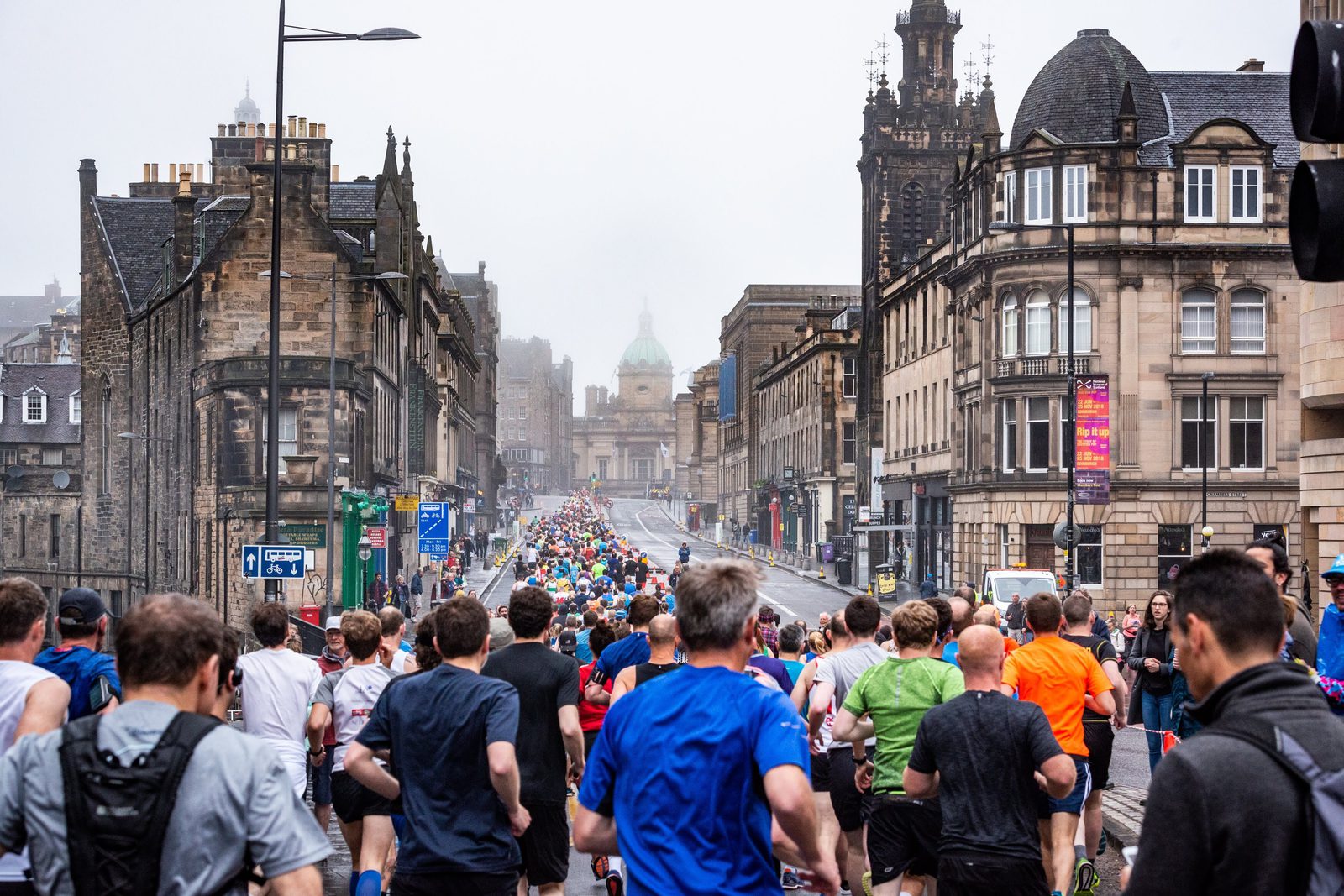 A large group of runners participating in a marathon moves through a city street lined with historic buildings. The scene is lively with participants wearing a mix of vibrant sportswear. The weather appears overcast, with a slight fog in the air.