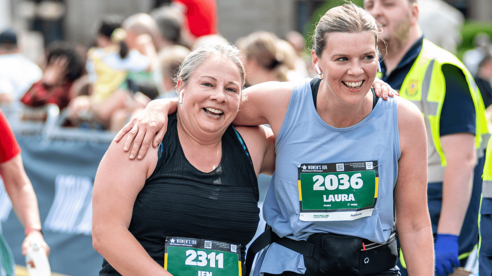 Two women in athletic gear, smiling and embracing after finishing a race. Both wear race bibs with the names Jen (2311) and Laura (2036). People and a medic are visible in the background.