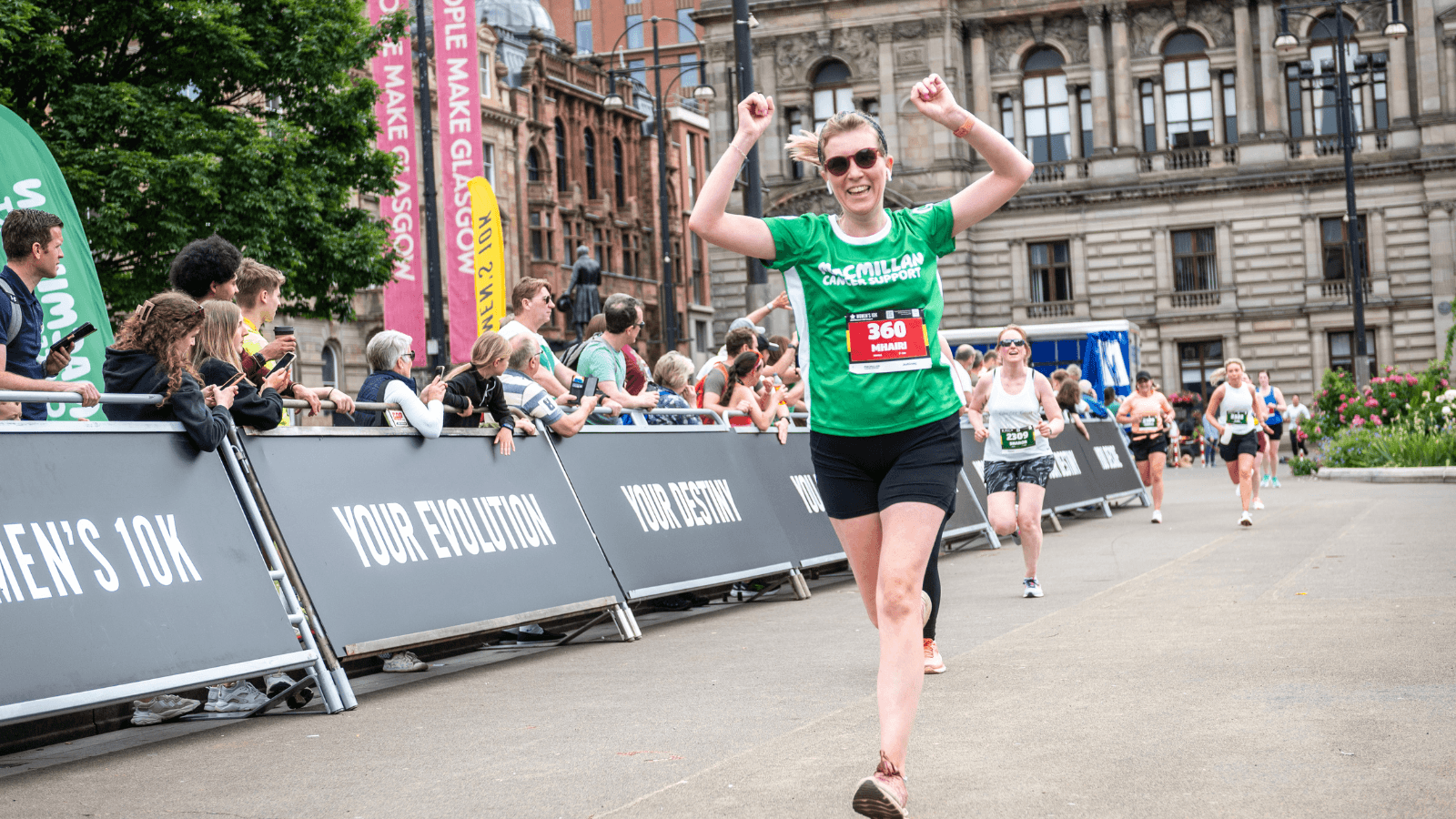 A woman in a green shirt and sunglasses smiles and raises her arms in celebration as she crosses the finish line of a road race, with spectators cheering behind barricades.