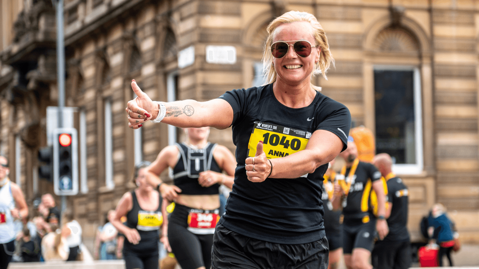 A smiling woman wearing sunglasses, a black t-shirt, and a race bib gives a thumbs-up while running in a city marathon; other runners and spectators are visible in the background.