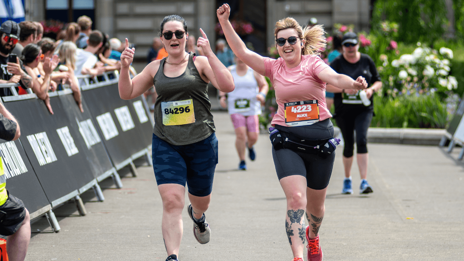 Two women are running toward the finish line in a race, cheering with their arms raised and smiling. Spectators line the sides, and other runners follow behind them on a sunny day.