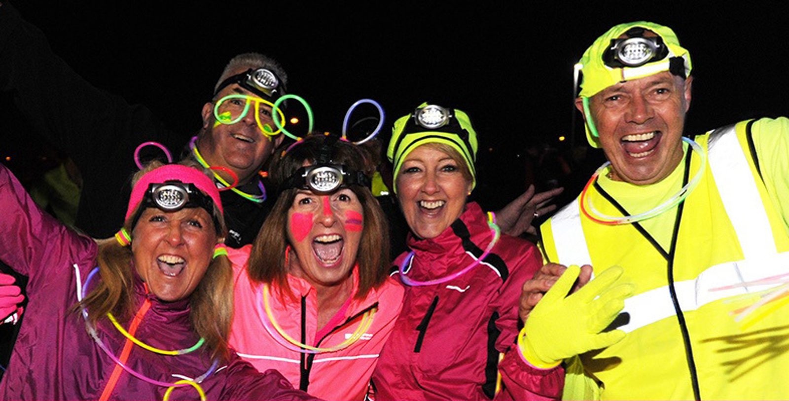 A group of five people wearing headlamps and neon clothing smiles and poses enthusiastically at a nighttime event. They are adorned with glow sticks and bright face paint against a dark background.