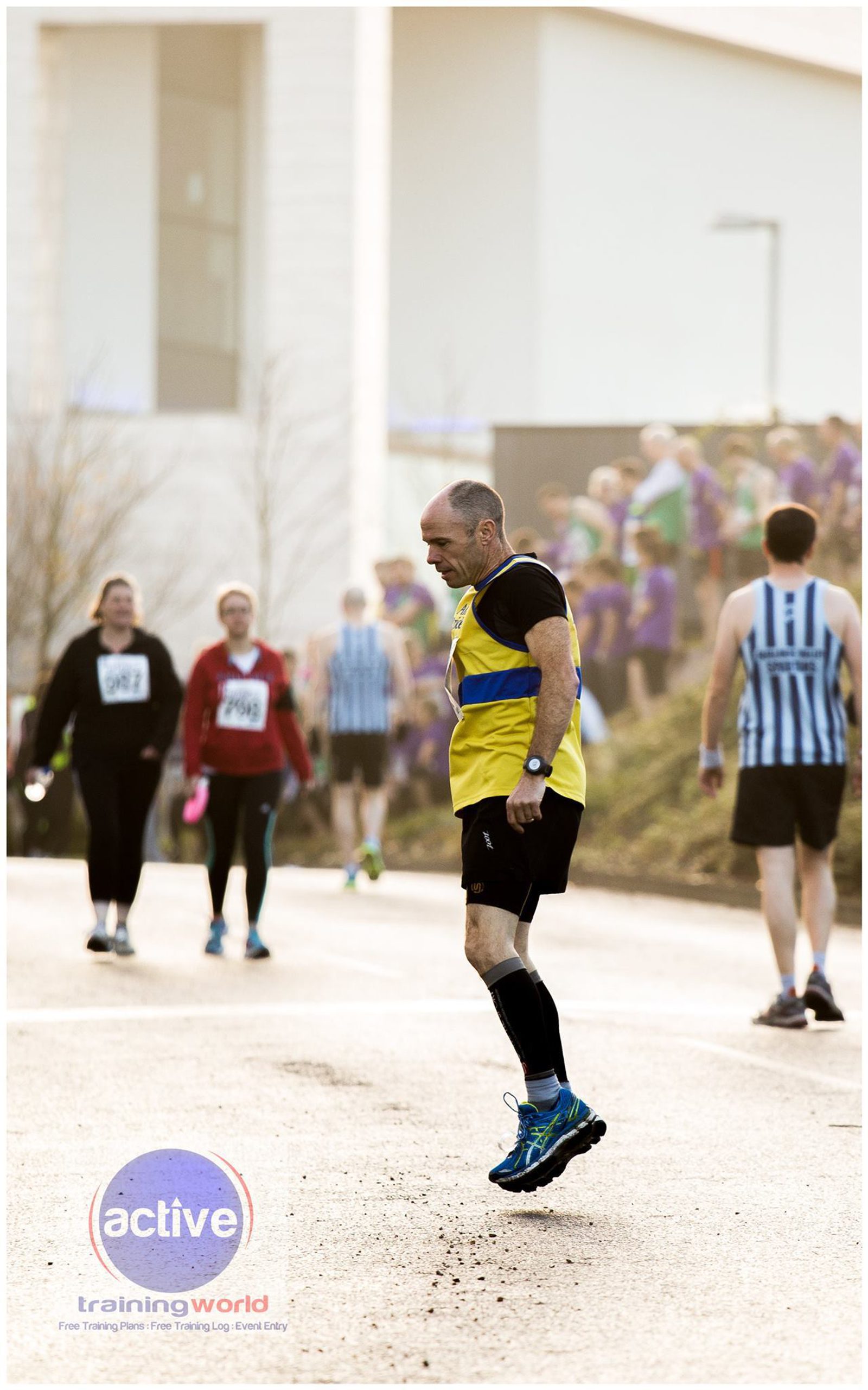 A runner in a yellow and black shirt is mid-stride on a road during a daytime race. Other participants and spectators are in the background, some wearing numbered bibs.