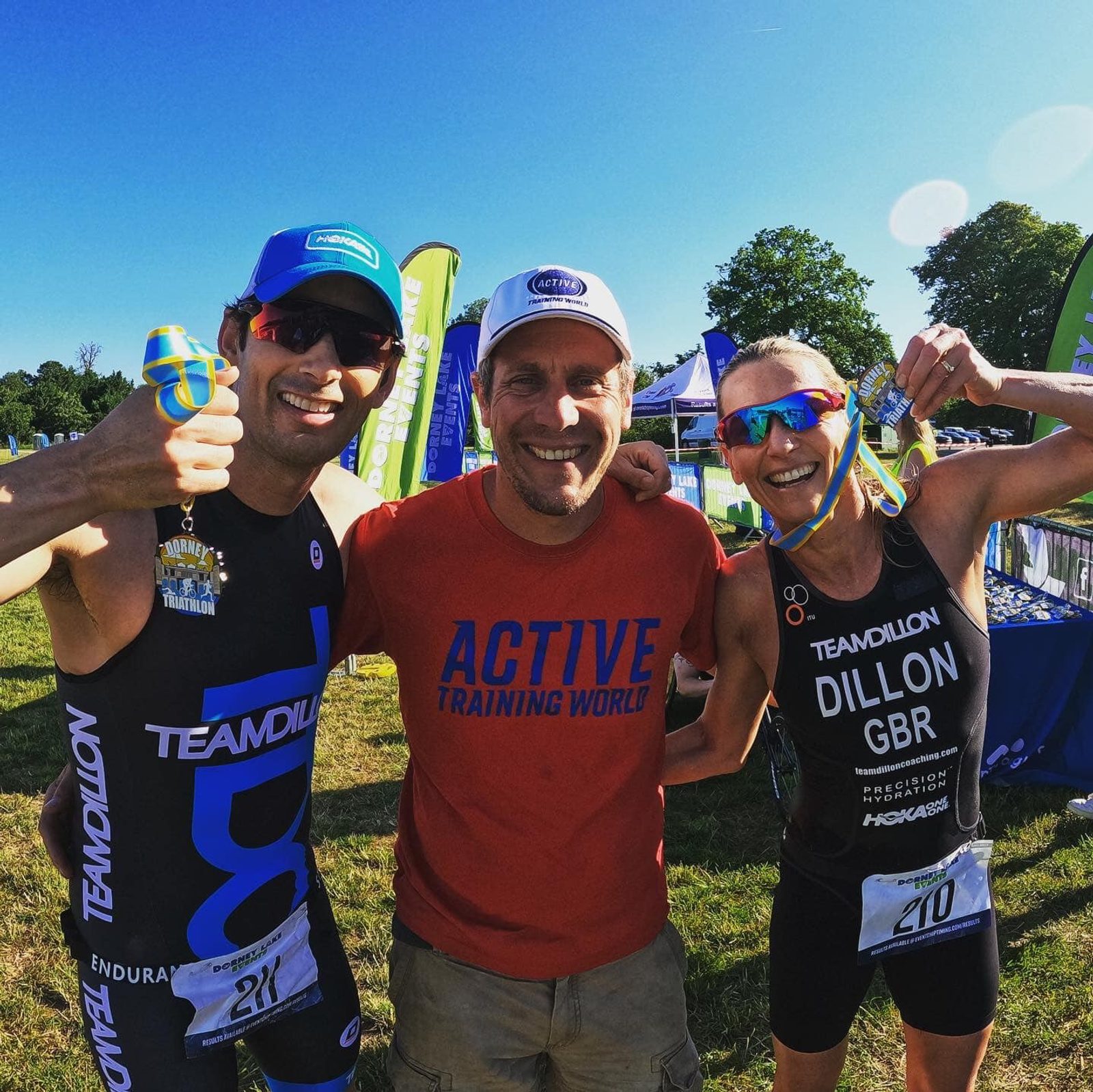 Three athletes celebrate at a triathlon event. The two on the left and right are wearing medals and athletic gear labeled "TEAM DILLON." The person in the middle wears a shirt reading "ACTIVE TRAINING WORLD." They're smiling, with green trees in the background.