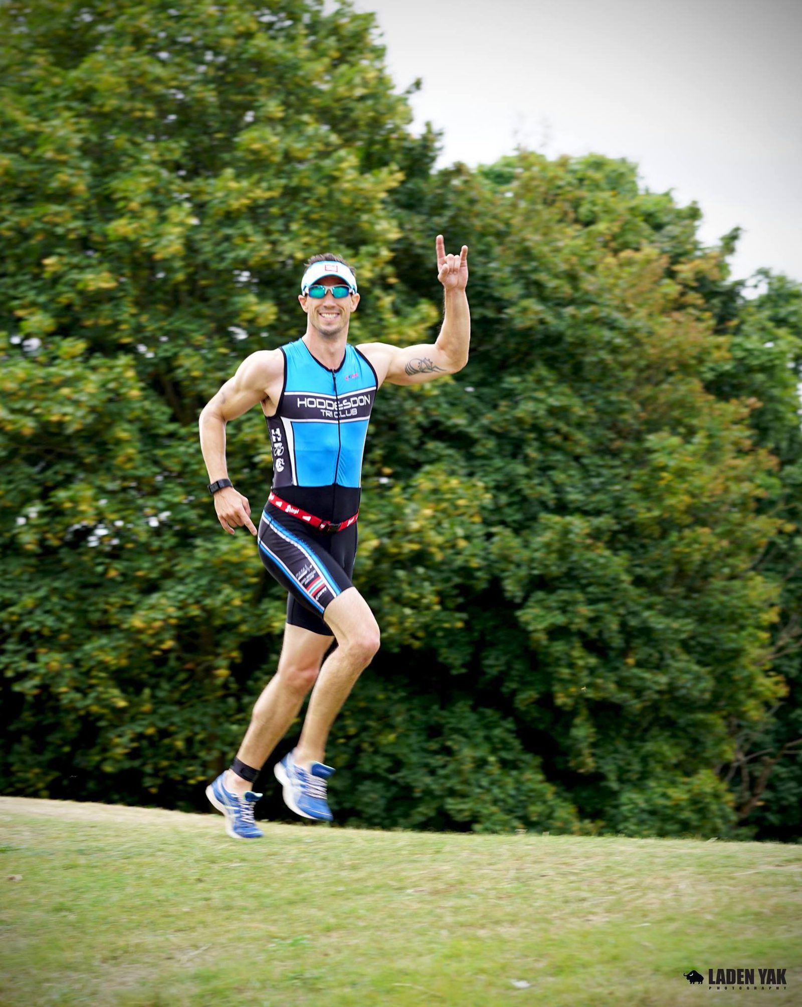 A man in a blue and black triathlon suit runs on grass, smiling and making a hand gesture. He wears sunglasses, a cap, and sneakers. Lush green trees are in the background under a cloudy sky.