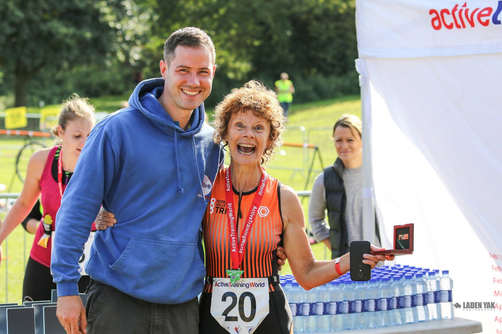 A smiling woman wearing a triathlon outfit and medal, holding an award next to a man in a blue hoodie. They're standing in a sunny outdoor event area near a table with water bottles. Other people can be seen in the background.