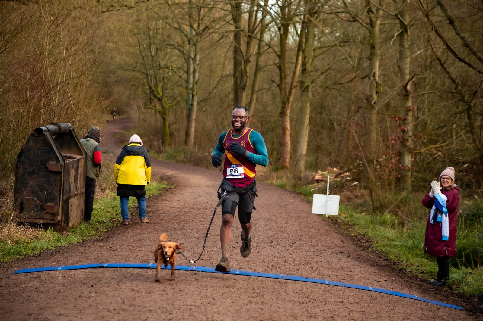 A man joyfully finishes a forest trail race with a small brown dog on a leash. He wears a colorful shirt and race bib. Two spectators cheer, standing nearby on the dirt path surrounded by trees.