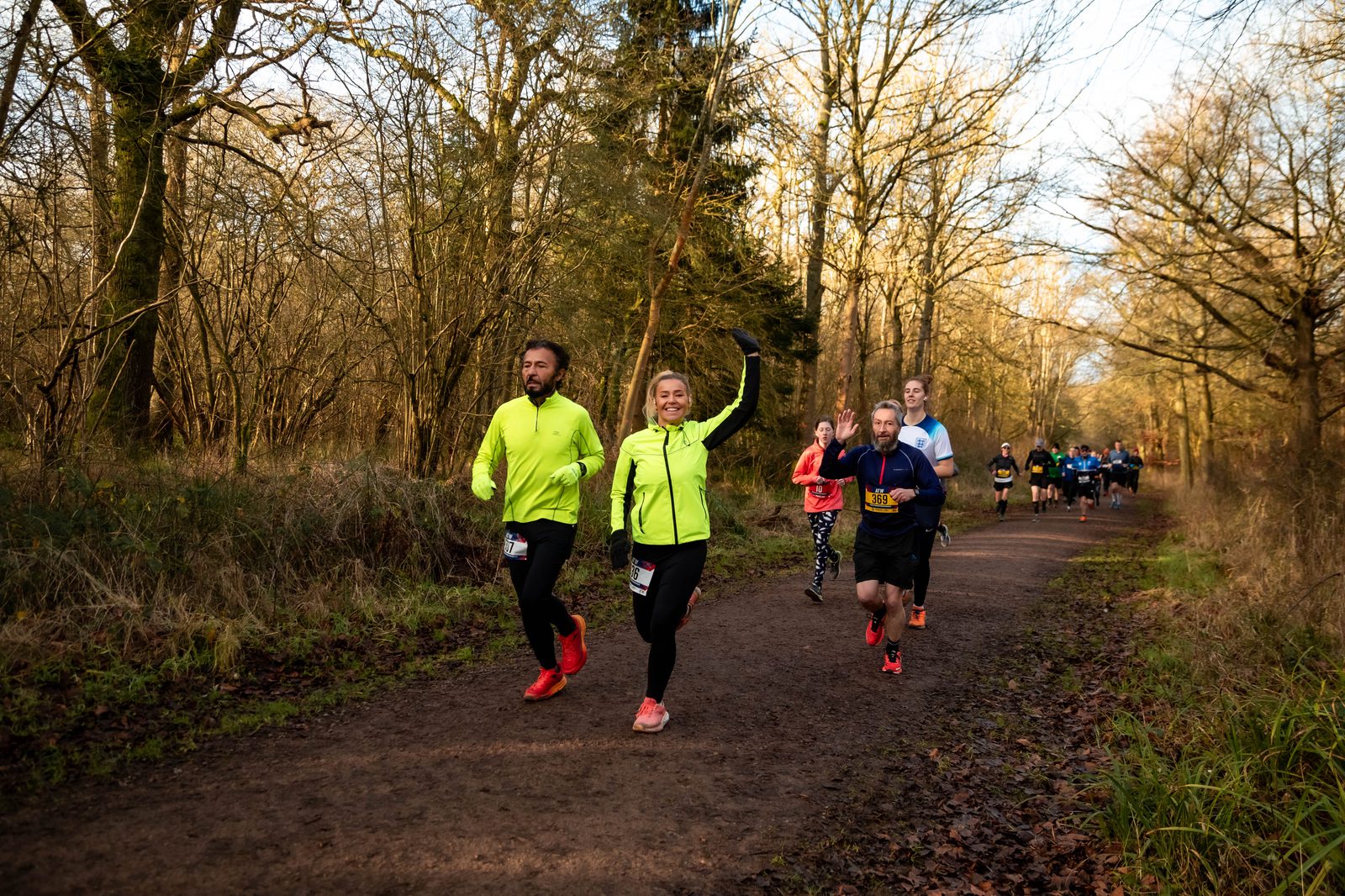 A group of runners jog along a wooded path on a sunny day. The front runners wear bright yellow jackets and smile, with others following behind. The trail is surrounded by trees and leaves.