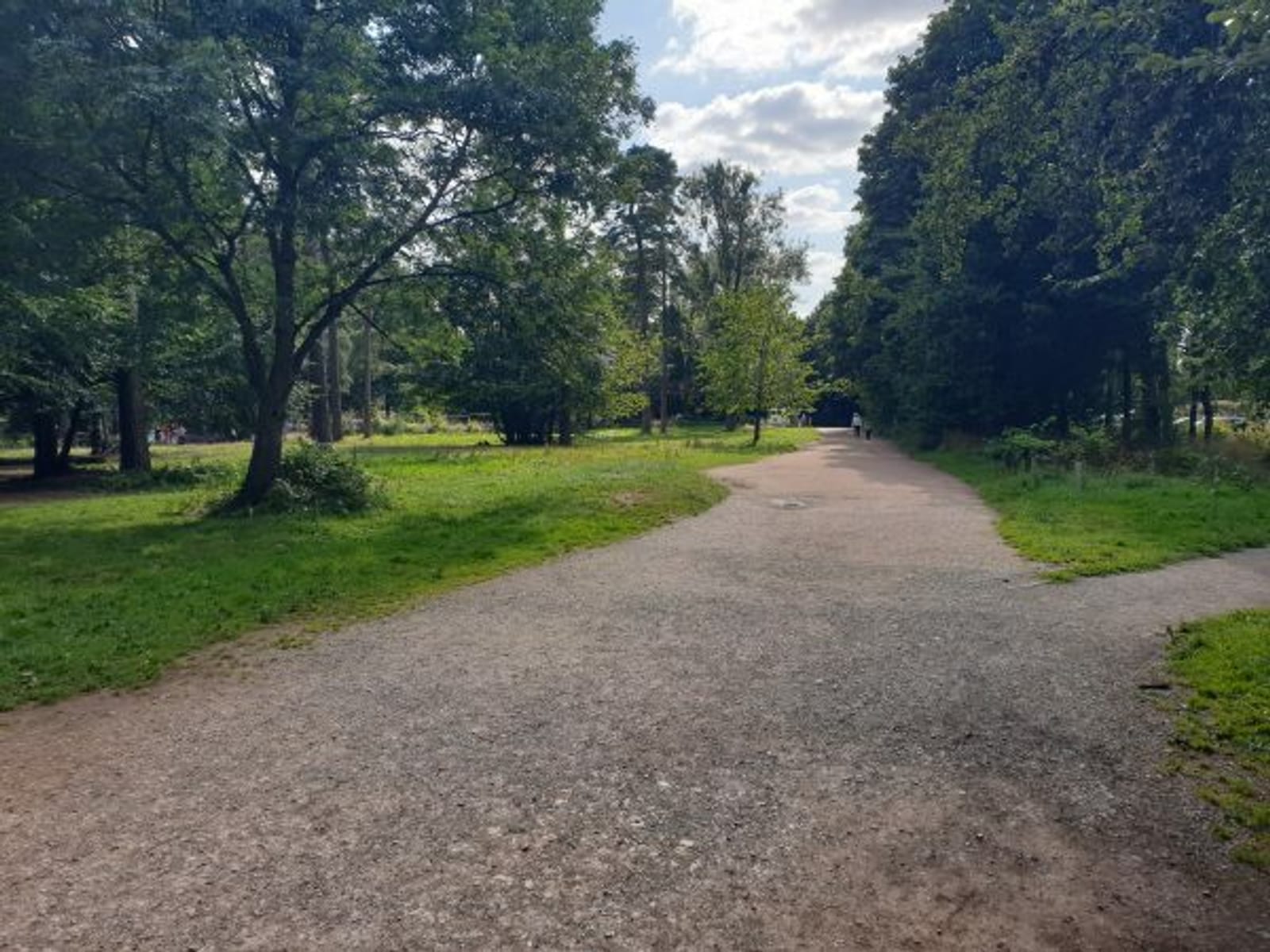A paved path winds through a lush park, surrounded by tall trees and greenery on either side. The sky is partly cloudy, casting a mix of light and shadow on the scene. The tranquil setting suggests a peaceful place for walking or enjoying nature.