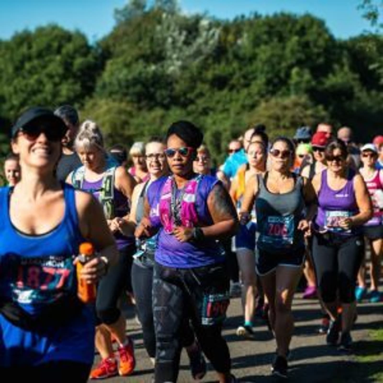 A diverse group of runners in athletic attire participate in an outdoor race, running along a path surrounded by greenery. The weather is sunny, and participants wear race bibs and some have medals. The atmosphere is energetic and competitive.