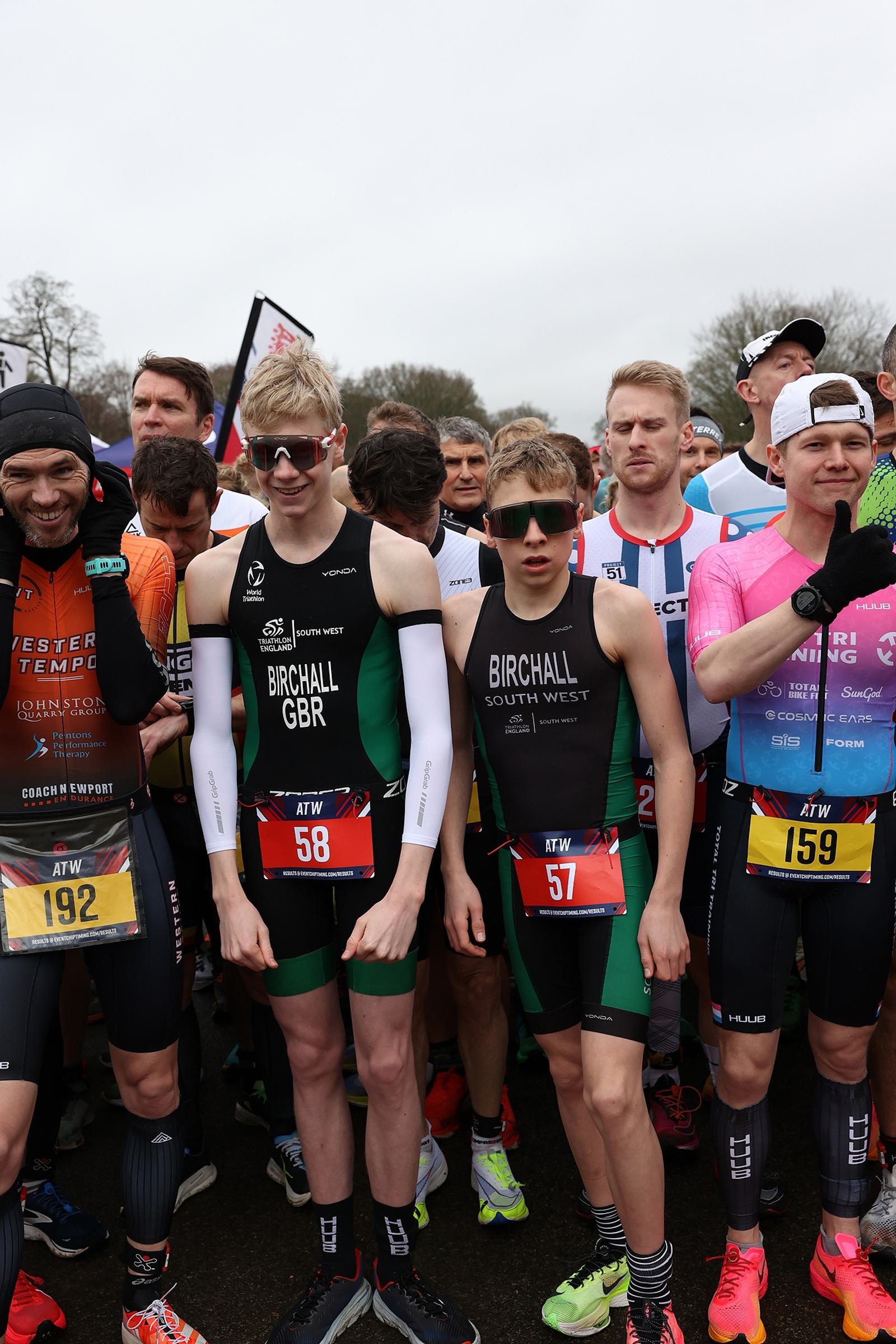 Athletes stand at the starting line of a race, wearing various sports gear and numbers. Some appear focused while others chat or smile. They are set against a backdrop of trees and a cloudy sky.