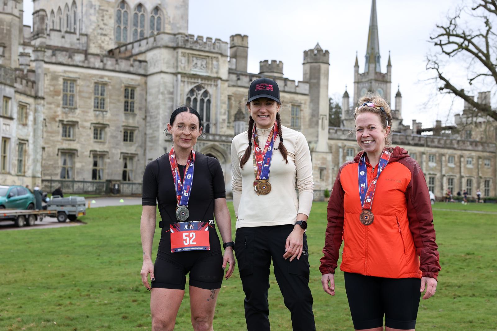 Three women stand in front of a historic building, each wearing medals and athletic clothing. One has a race bib numbered 52. It appears to be a post-race celebration. The setting is a grassy area with a castle-like structure in the background.