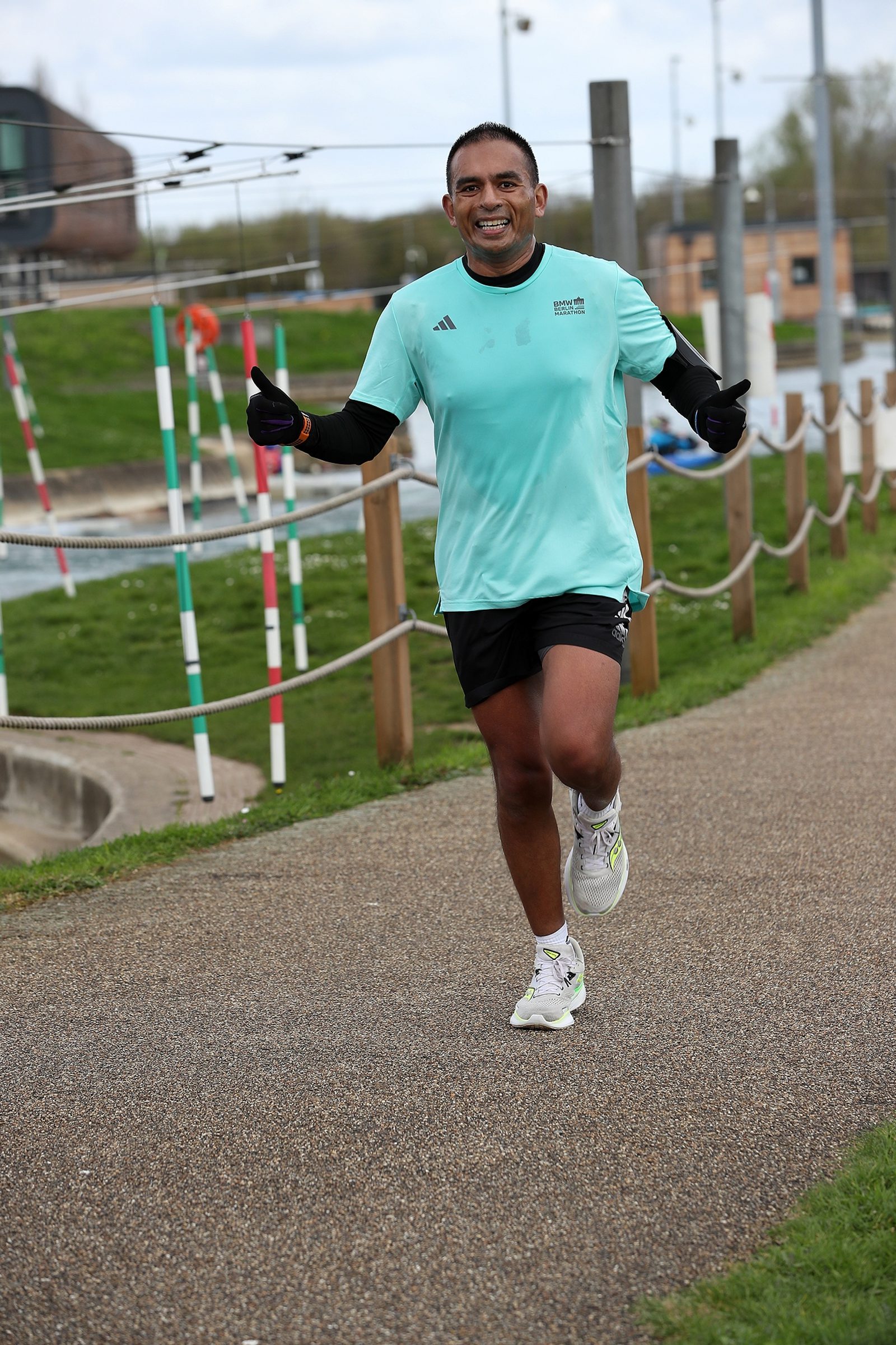 A man in a light blue shirt and black shorts is jogging on a park path. He is smiling, giving a thumbs up with both hands. The background features grass, poles, and a building.