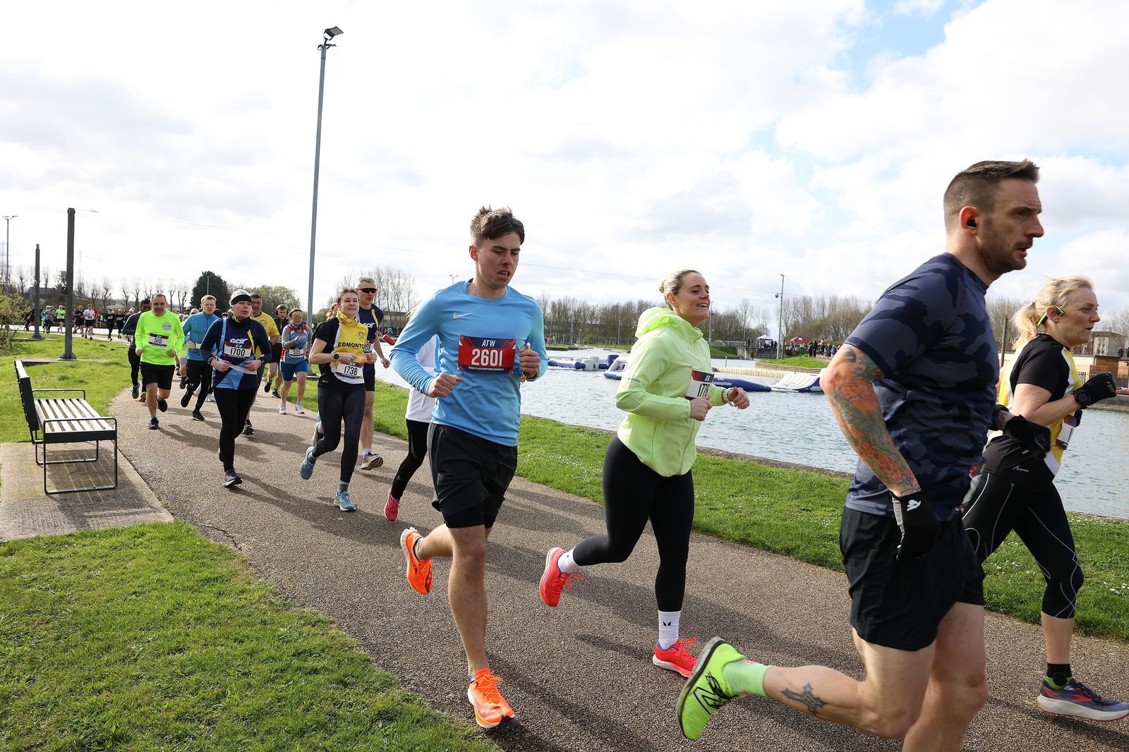 A group of people running along a riverside path during a race. The runners are dressed in athletic wear, and the background shows trees, grass, and a cloudy sky. It appears to be a lively outdoor event.