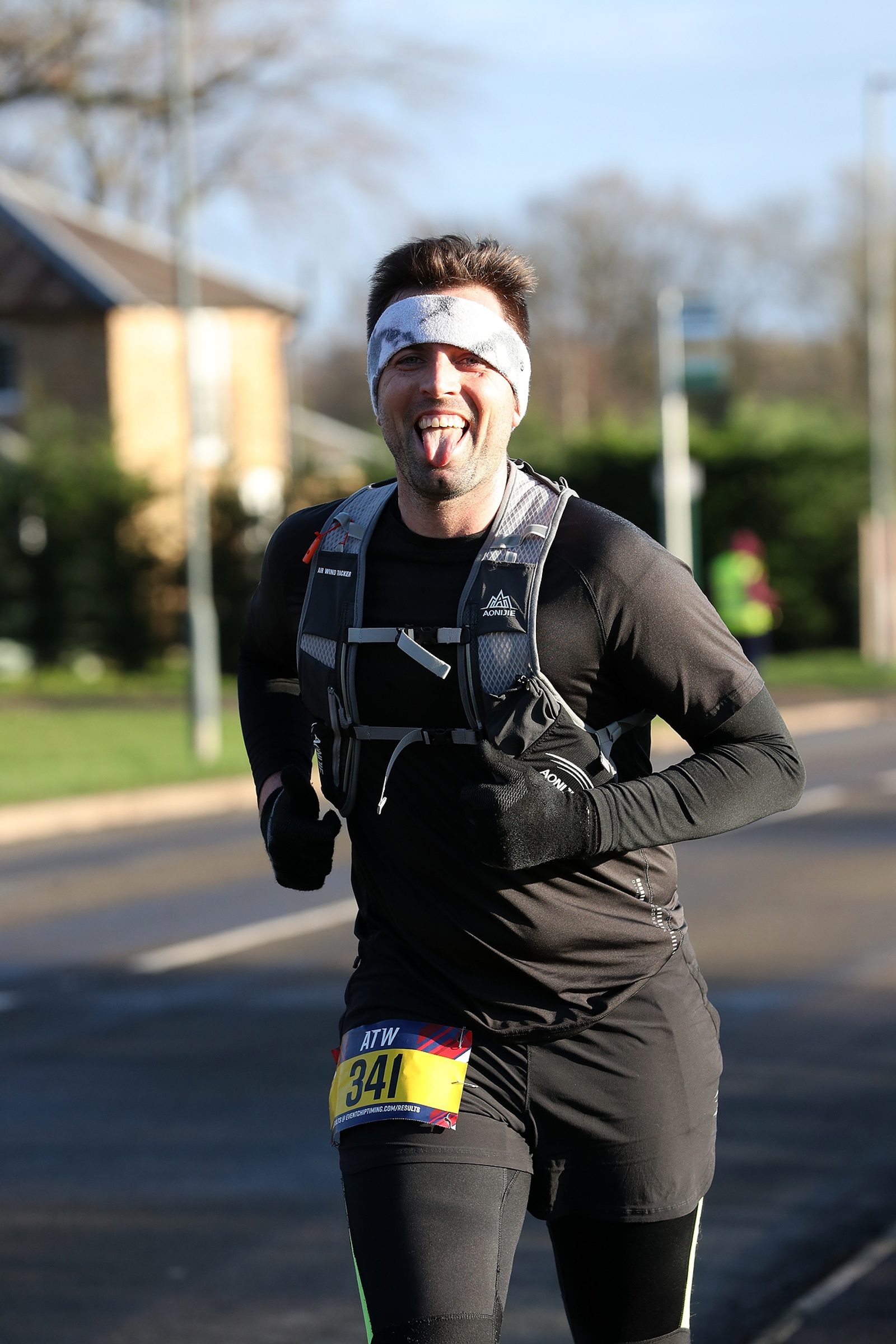 A male runner wearing a black outfit and white headband jogs on a sunny day, smiling with his tongue out. He has a race bib numbered 341 attached to his waist. Houses and greenery are visible in the background.