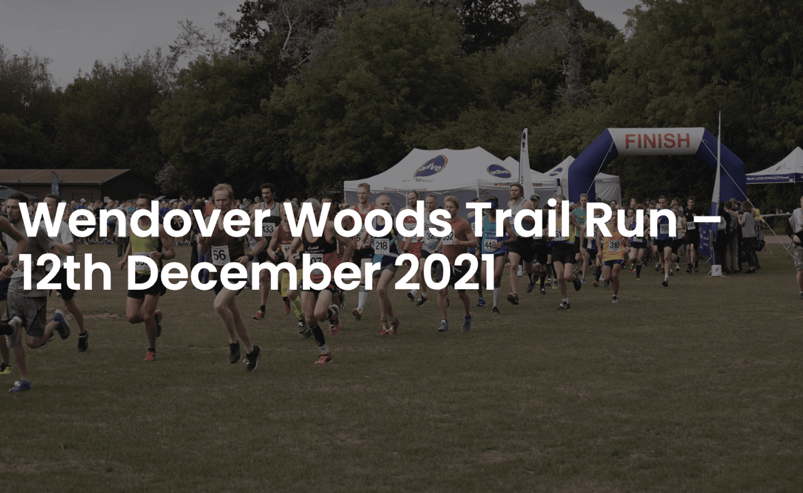 Runners start a race in an outdoor, wooded setting at the Wendover Woods Trail Run on December 12th, 2021. A large inflatable arch that says "FINISH" is in the background. Trees and event tents are visible.