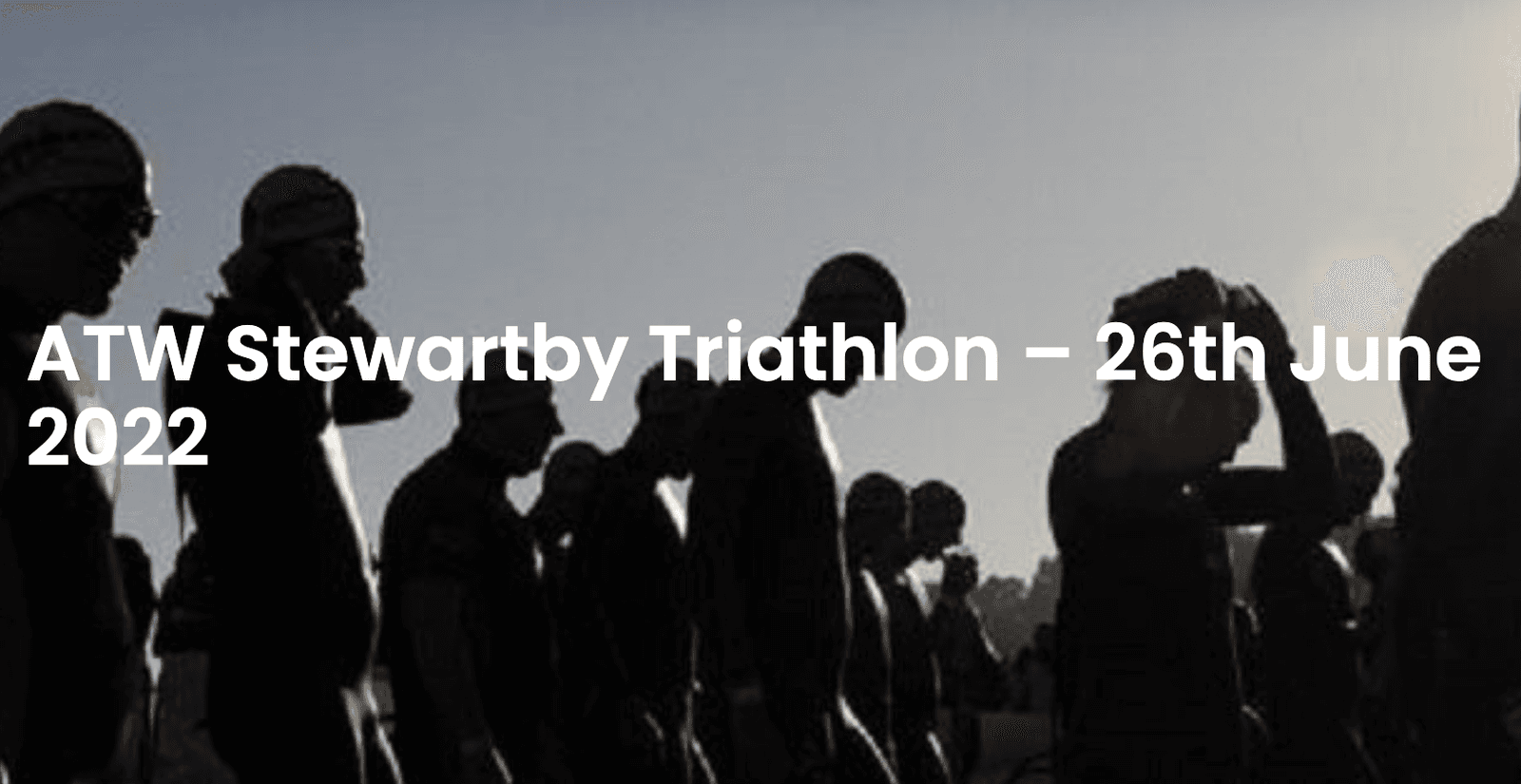 A group of triathletes in wetsuits and swim caps prepare to start a race at the ATW Stewartby Triathlon on June 26, 2022. The sun is low in the sky, casting the athletes in silhouette.
