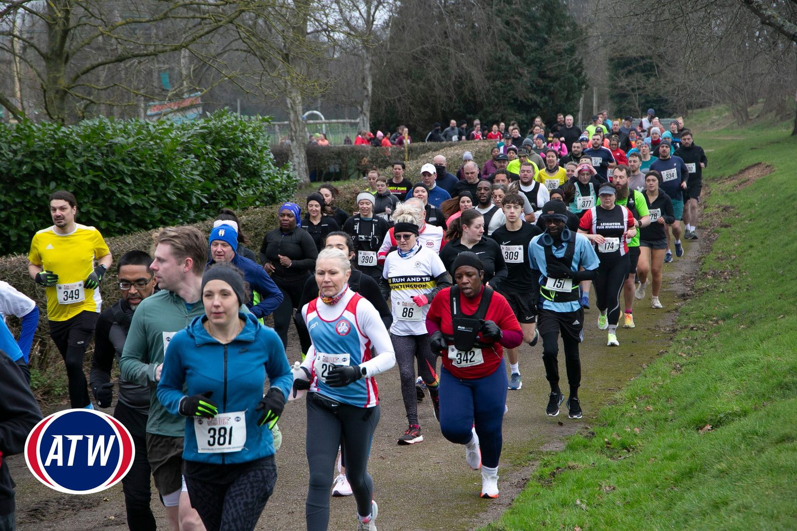 A large group of runners, dressed in athletic gear and numbered bibs, participate in a race on a park path surrounded by trees and greenery. An ATW logo is in the lower left corner.