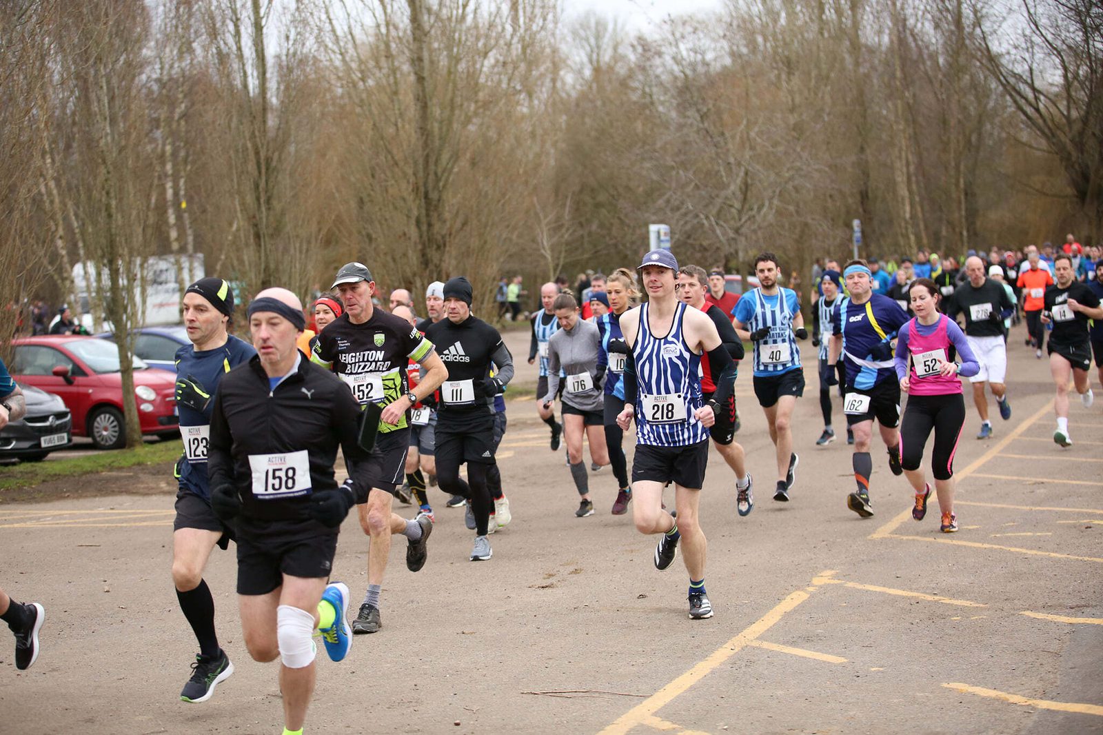 A group of runners in winter athletic gear participate in a road race on a cold day, with barren trees and parked cars in the background. Each runner wears a numbered bib, with some focused on the path ahead while others acknowledge the environment around them.