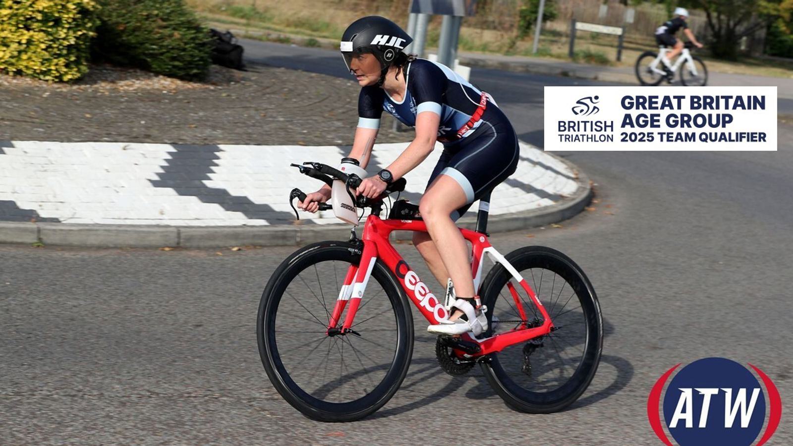 A cyclist is participating in a race, wearing a blue and white cycling uniform and helmet. They are riding a red and white bicycle. In the background, there is a sign reading "Great Britain Age Group 2025 Team Qualifier" with British Triathlon and ATW logos.