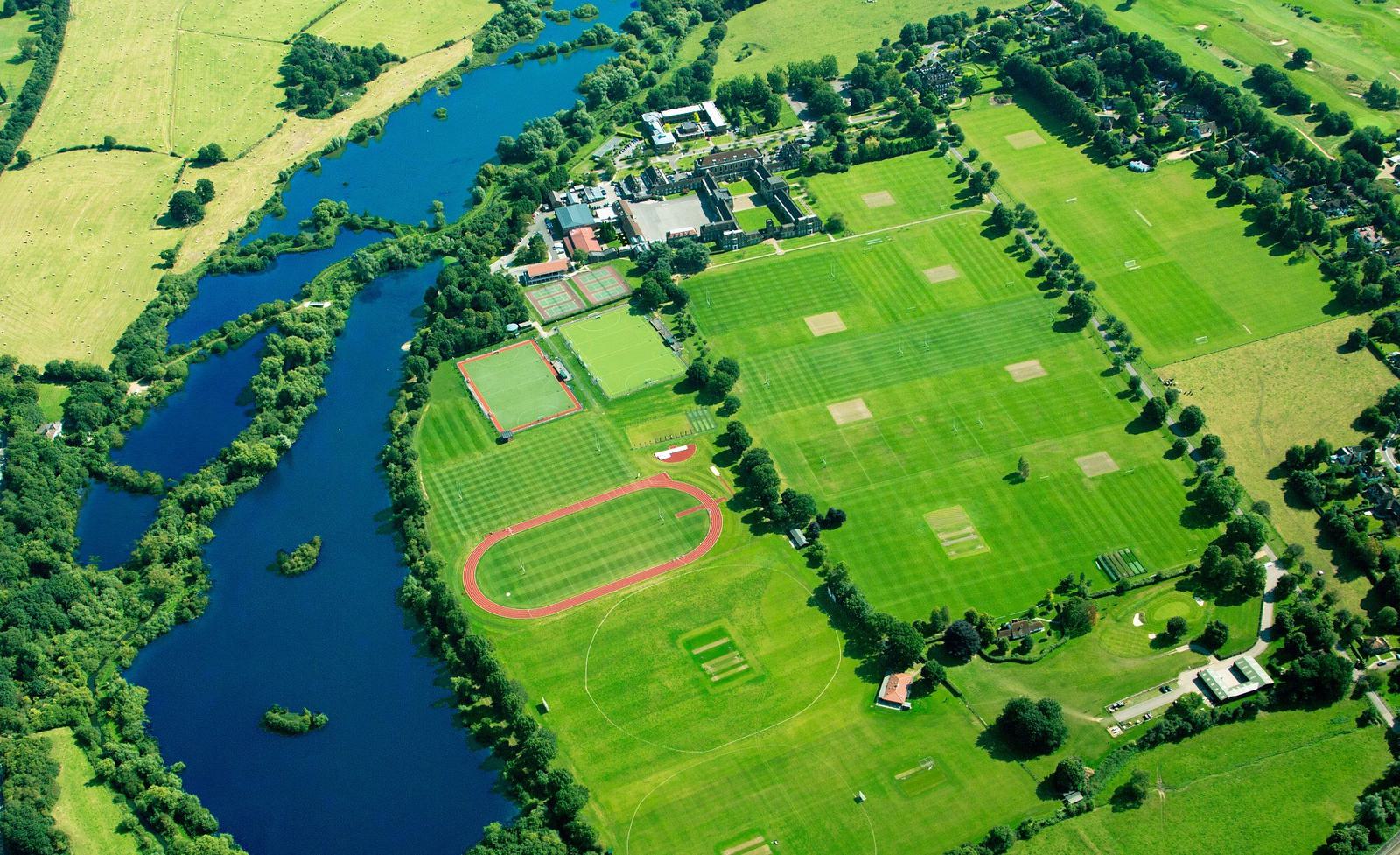 Aerial view of a large green sports complex with multiple fields, a running track, tennis courts, buildings, and a nearby river. Surrounding areas include trees, roads, and open green spaces. The complex is surrounded by rural landscapes.