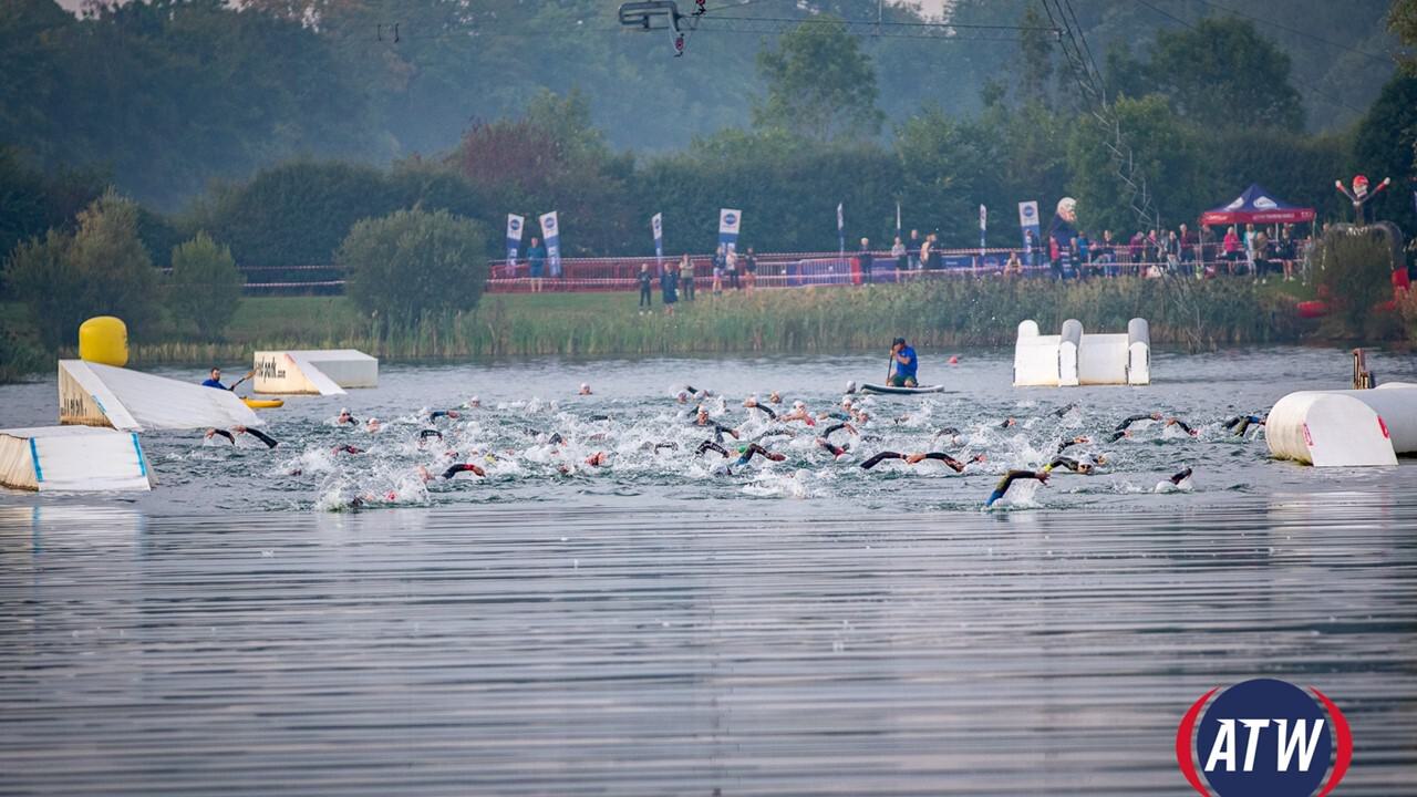 Triathletes swimming in a lake during a race, with buoys and floating barriers marking the course. A referee on a boat oversees the participants. Spectators can be seen on the shore under tents. The ATW logo is visible in the lower-right corner of the image.