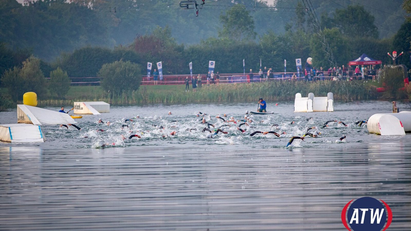 Triathletes swimming in a lake during a race, with buoys and floating barriers marking the course. A referee on a boat oversees the participants. Spectators can be seen on the shore under tents. The ATW logo is visible in the lower-right corner of the image.