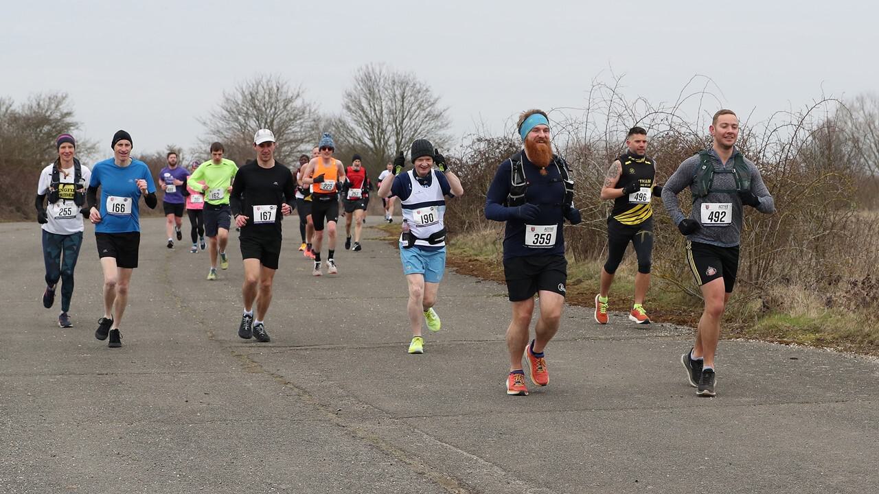 A group of runners on a paved path during an outdoor race. Participants are wearing various athletic outfits, bib numbers, and some have hydration packs. Trees without leaves and overcast sky in the background suggest a chilly day.