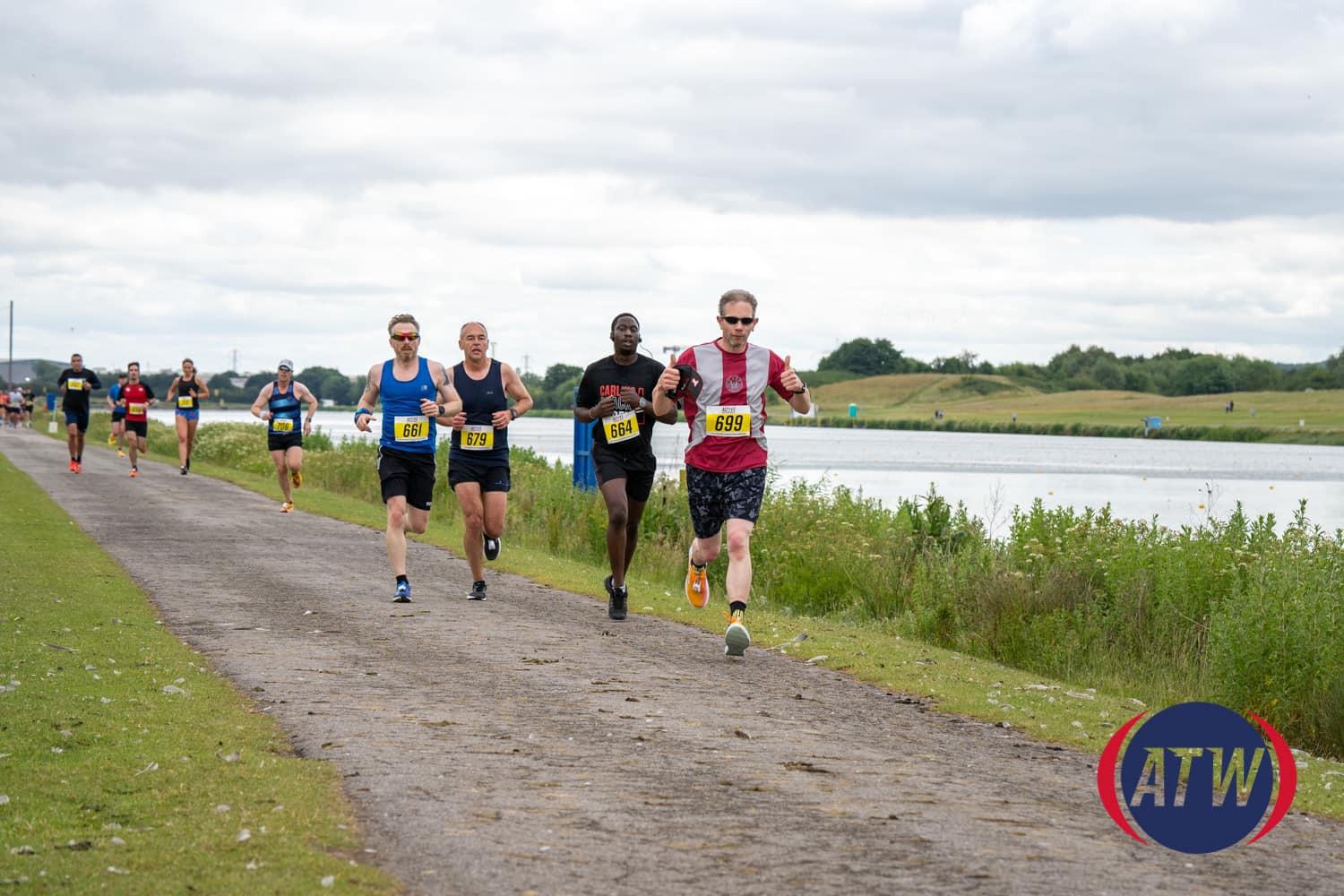 A group of runners participates in a race on a paved path alongside a large body of water. The sky is overcast, and the landscape is green and open. The runners are wearing numbered bibs and athletic gear. A logo with "ATW" is visible in the bottom right corner.