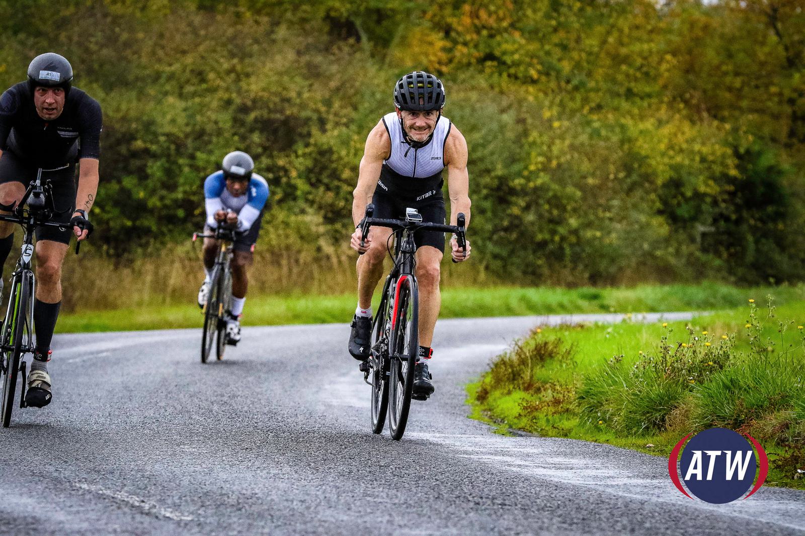 Three cyclists are riding on a winding road with trees and greenery in the background. They are wearing helmets and professional cycling attire. The cyclist in the foreground appears to be leading, with two cyclists following closely behind. The ATW logo is visible in the corner.