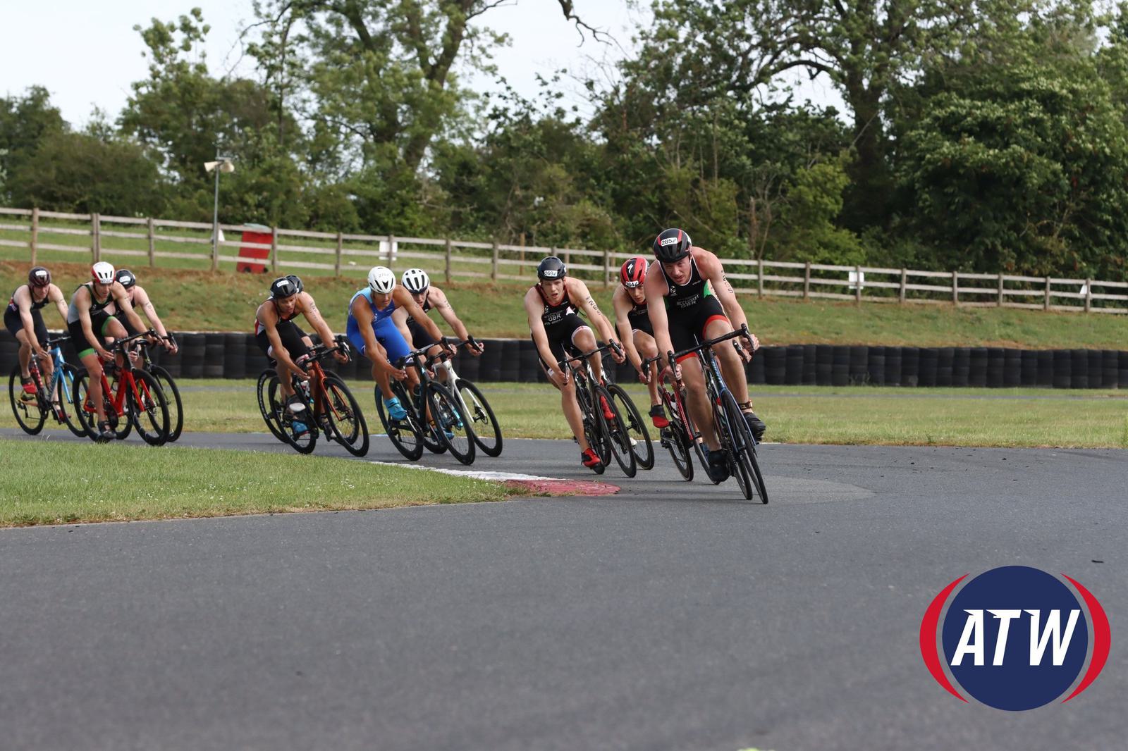 A group of cyclists tightly packed together racing on a paved road in an outdoor setting with greenery and trees in the background. The cyclist in the lead is leaning into a curve. The ATW logo is visible in the bottom right corner.