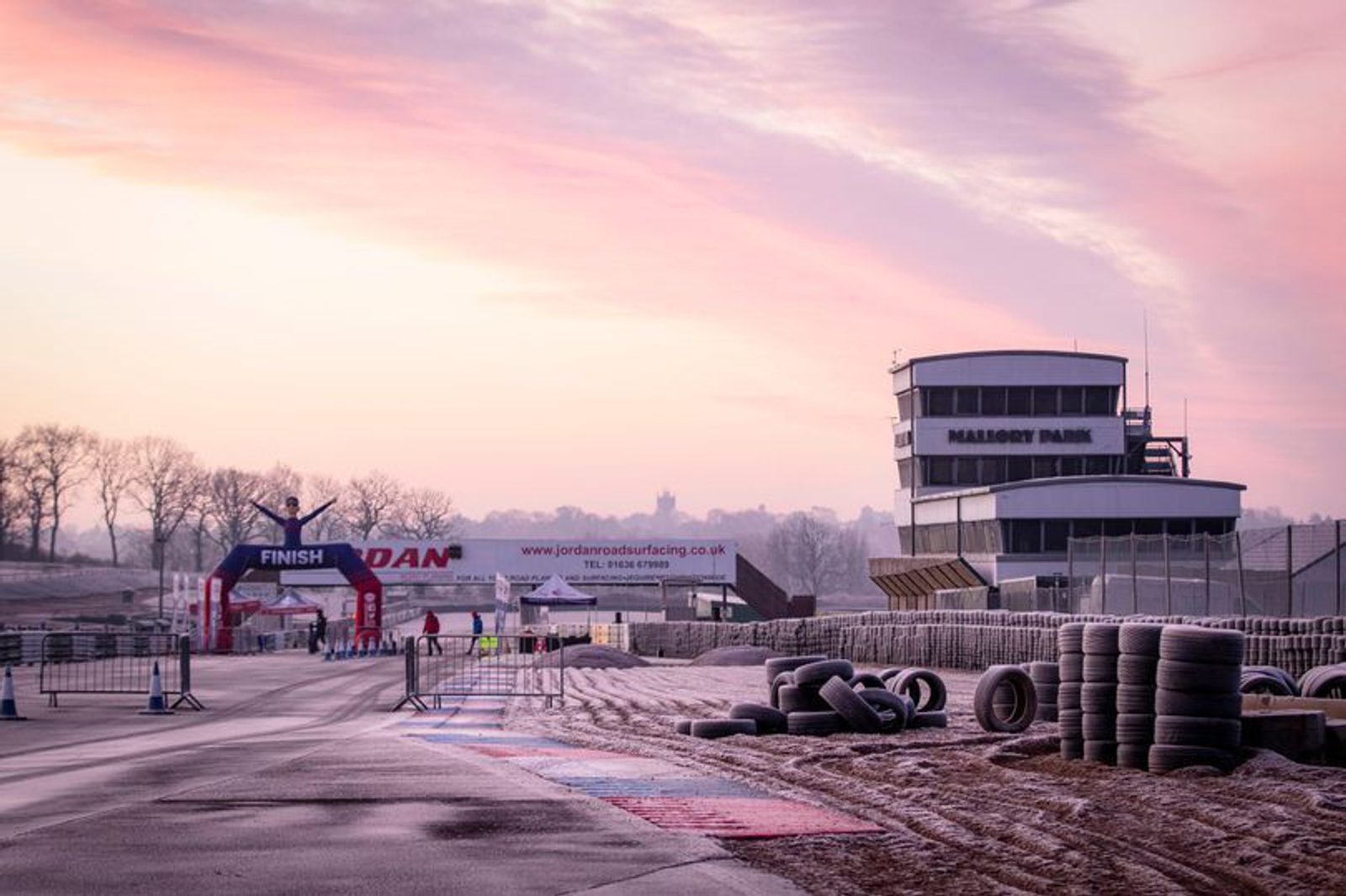 A race track at dawn with a visible "FINISH" inflatable archway and a building labeled "MALLORY PARK." Tire barriers line the track, and a few people stand near the finish area. The sky has soft pastel colors, adding a serene atmosphere.
