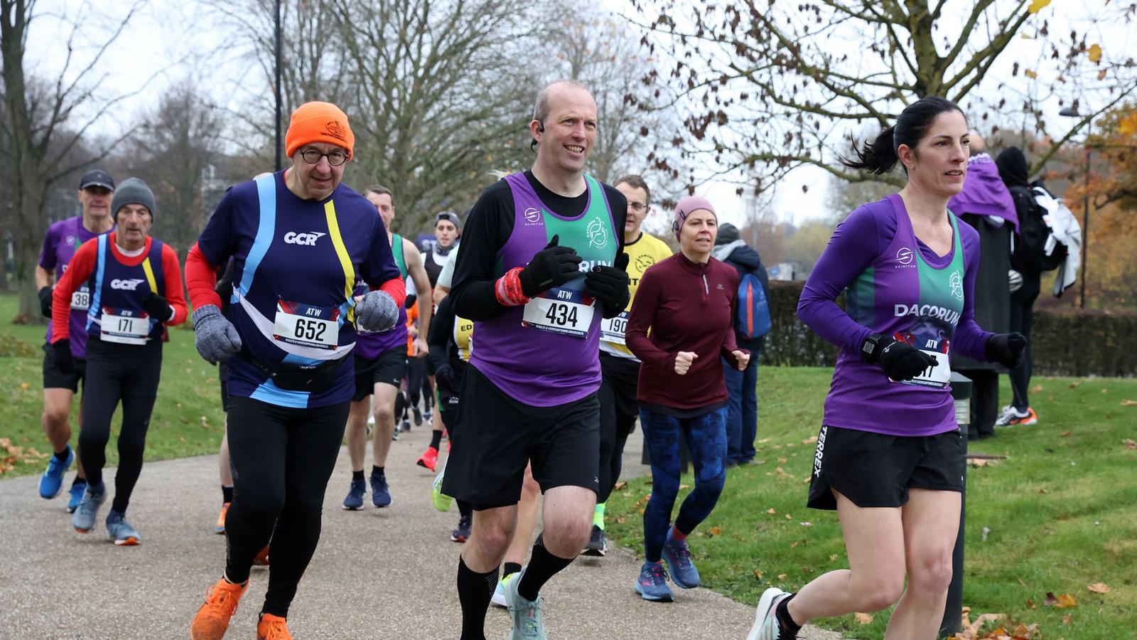 A group of runners participating in a race, wearing various colored running gear and numbered bibs. They are running on a paved path in a park setting with trees and autumn leaves in the background. The atmosphere is lively and energetic.