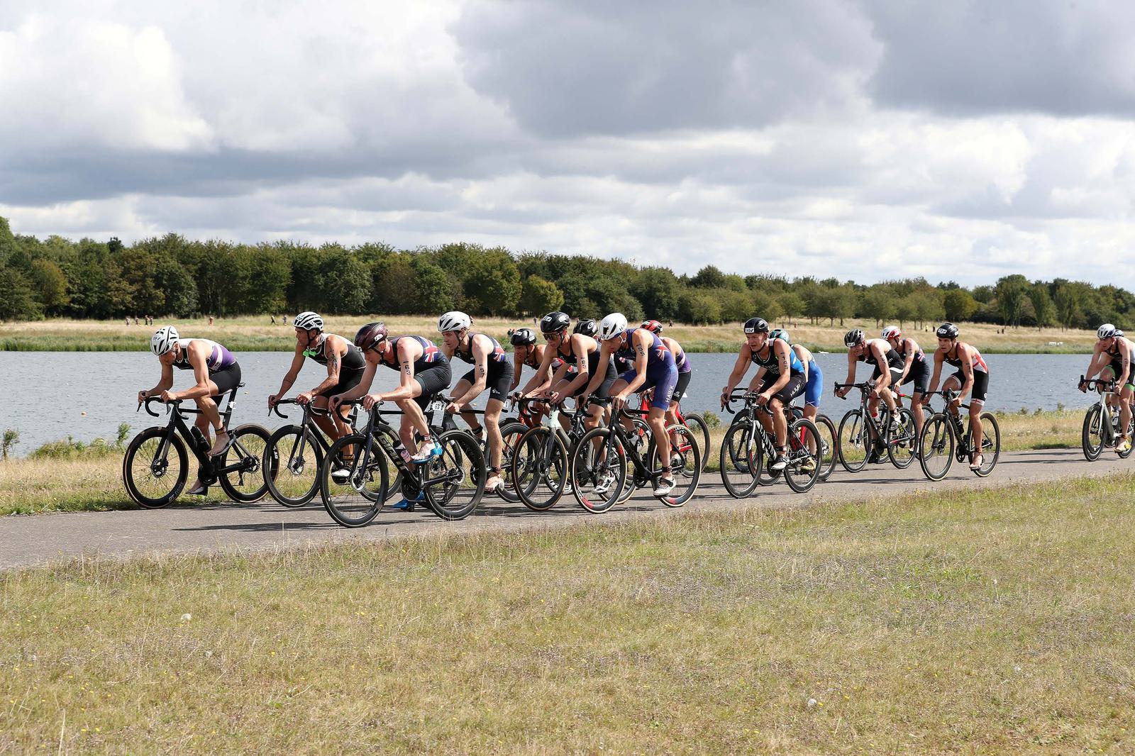 A group of cyclists wearing helmets race along a paved road beside a body of water. The landscape is grassy with dense trees in the background. The sky is overcast with scattered clouds.