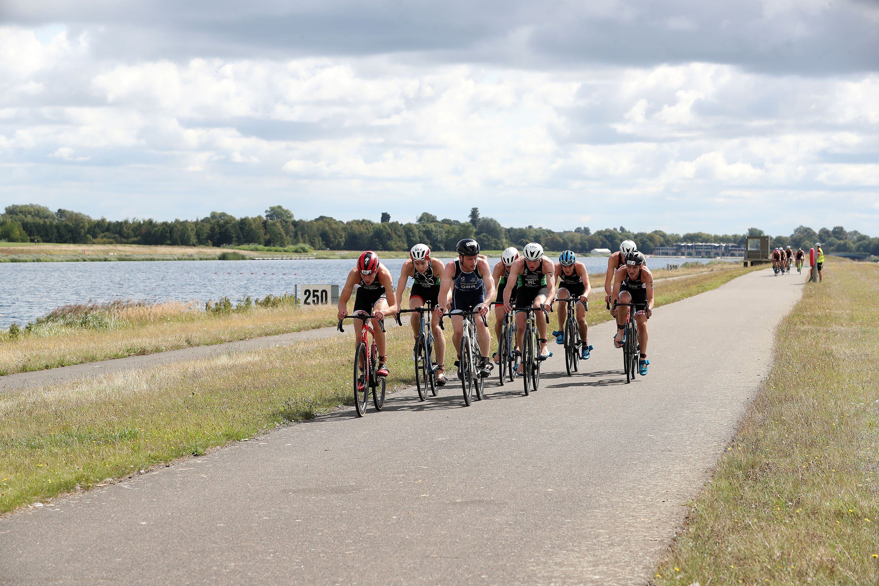 A group of cyclists rides along a paved path beside a body of water under a partly cloudy sky. The road has grass on both sides, and a sign on the left reads "250m." More cyclists can be seen in the distance.
